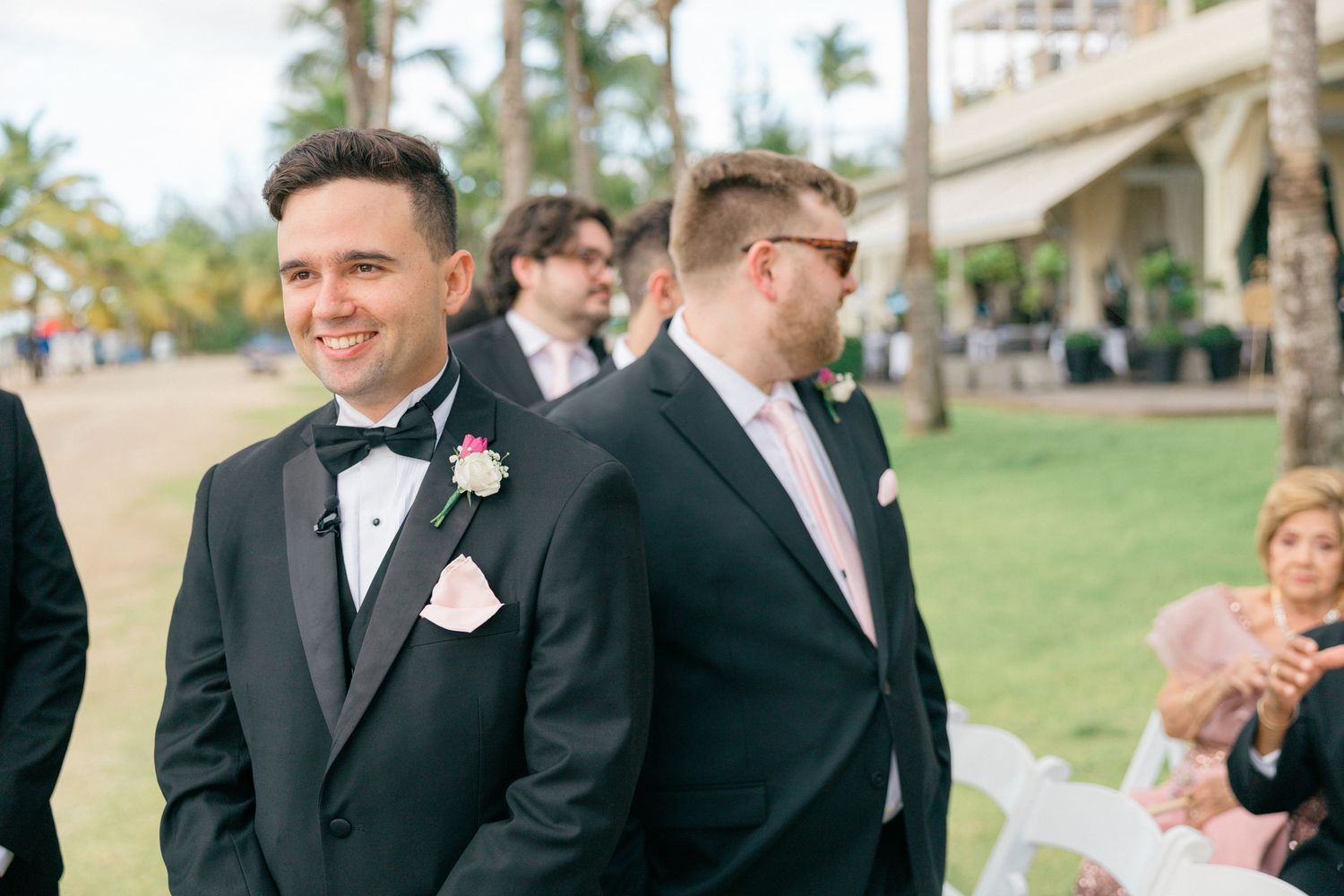 Groomsmen in black tuxedos stand together at an outdoor tropical wedding venue.