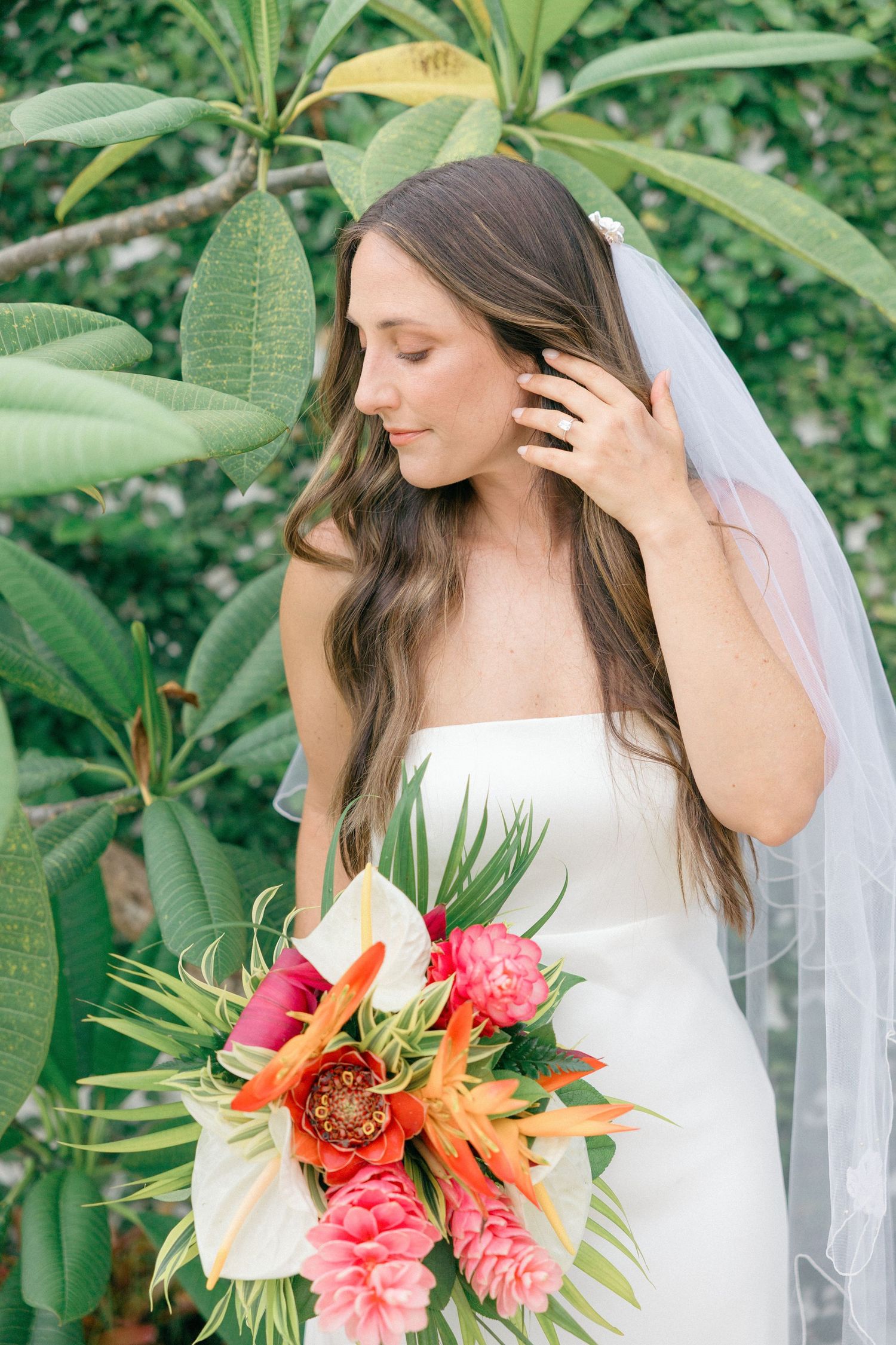 Bride holds vibrant tropical bouquet featuring orange lilies and pink blooms against green garden backdrop.