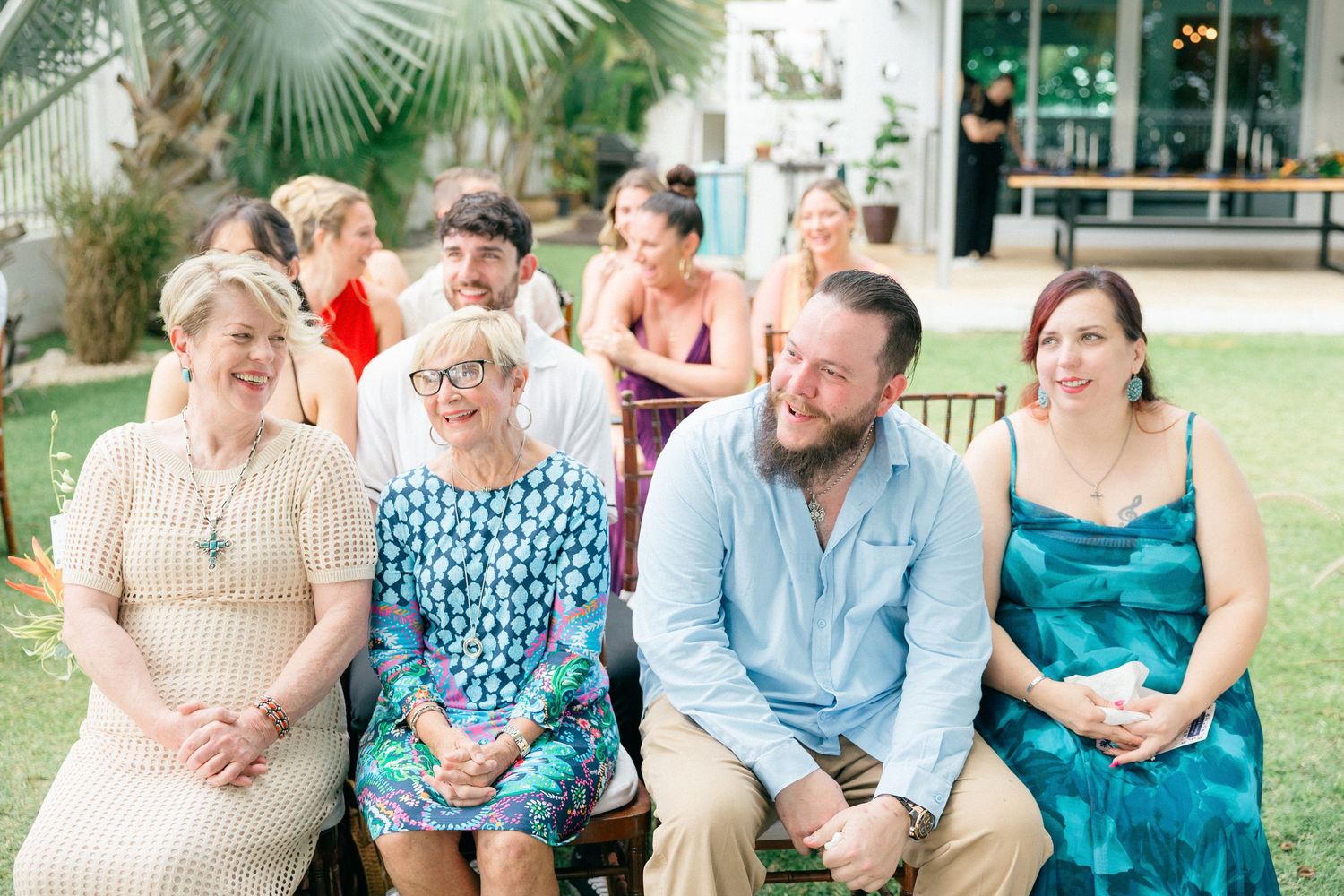 Guests in colorful summer attire enjoy outdoor wedding ceremony seating area.
