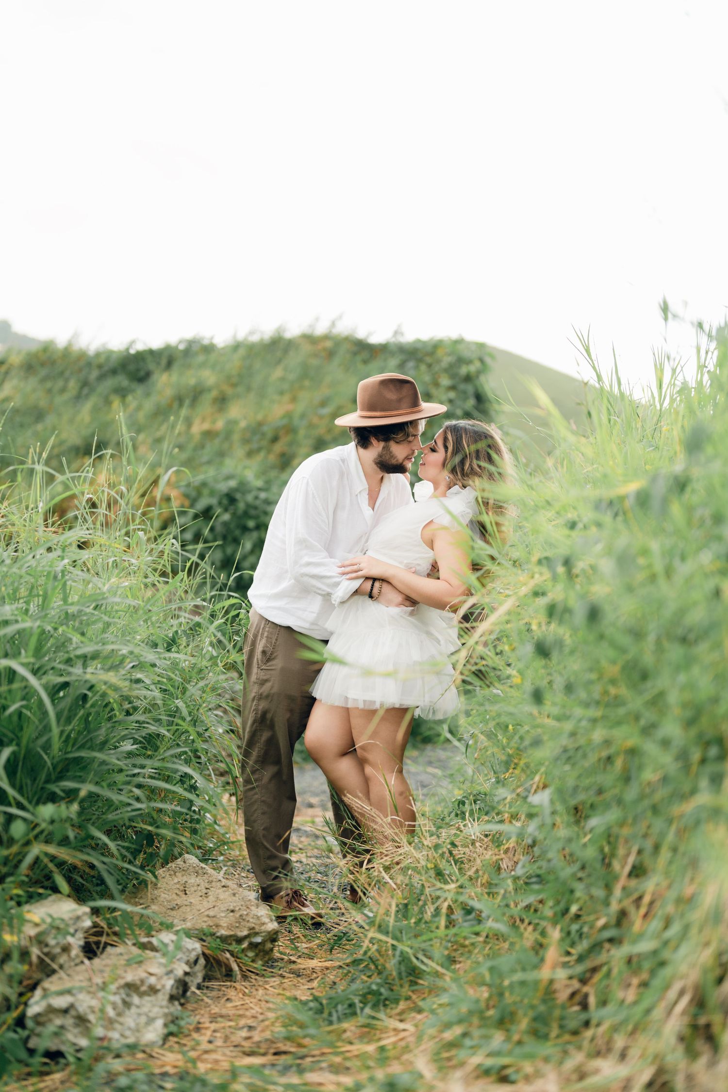 Romantic couple embracing on a hillside wearing white clothing and a vintage hat.