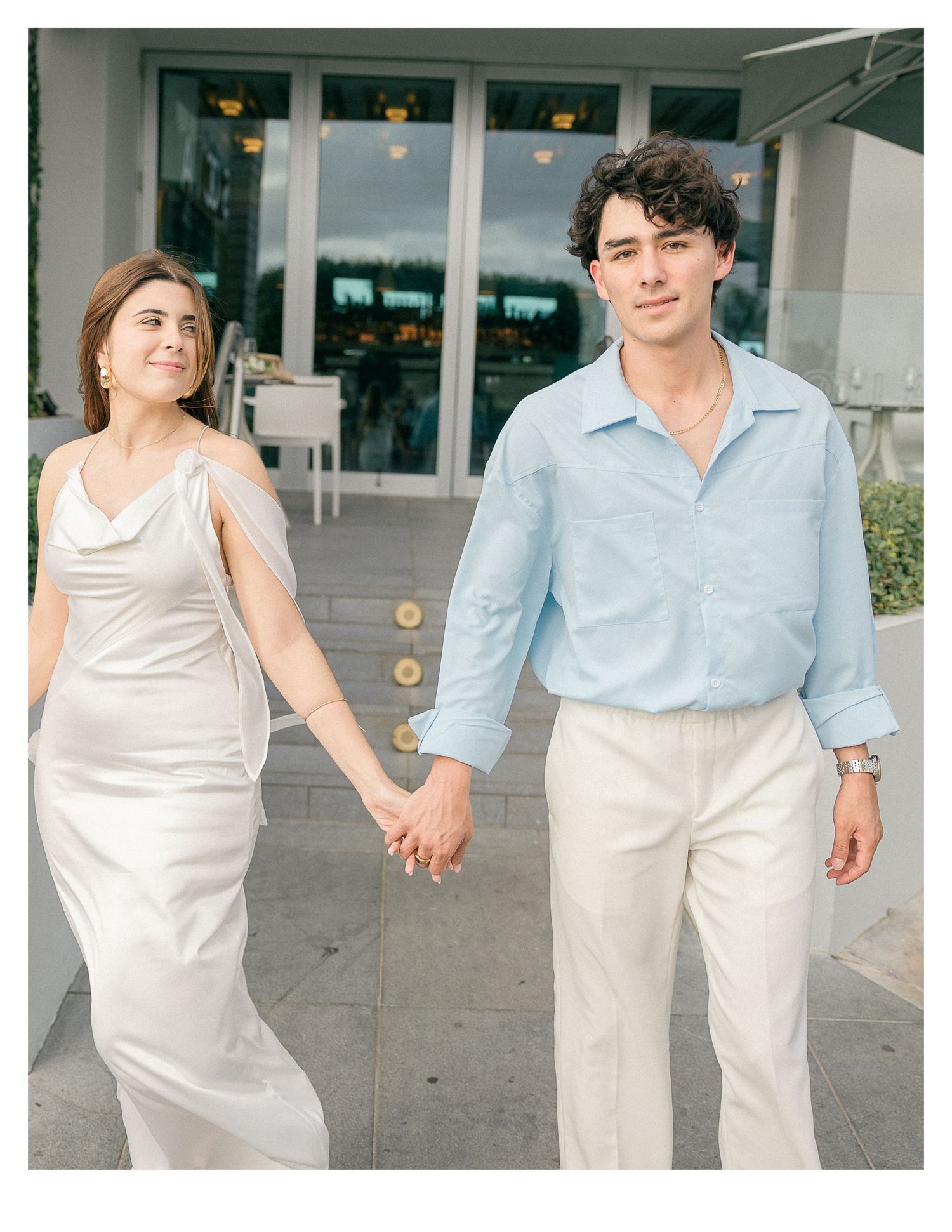 A couple in white and blue outfits walking together outside a modern building.