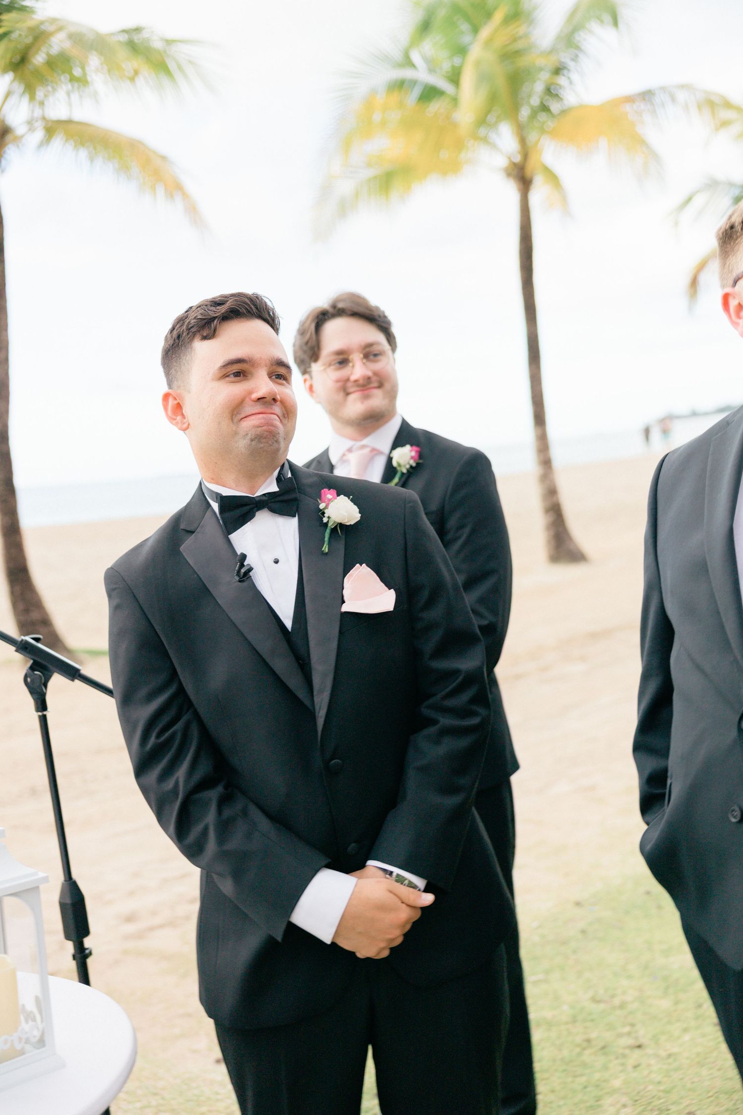 Groomsmen in black tuxedos stand together at a beachside wedding ceremony.