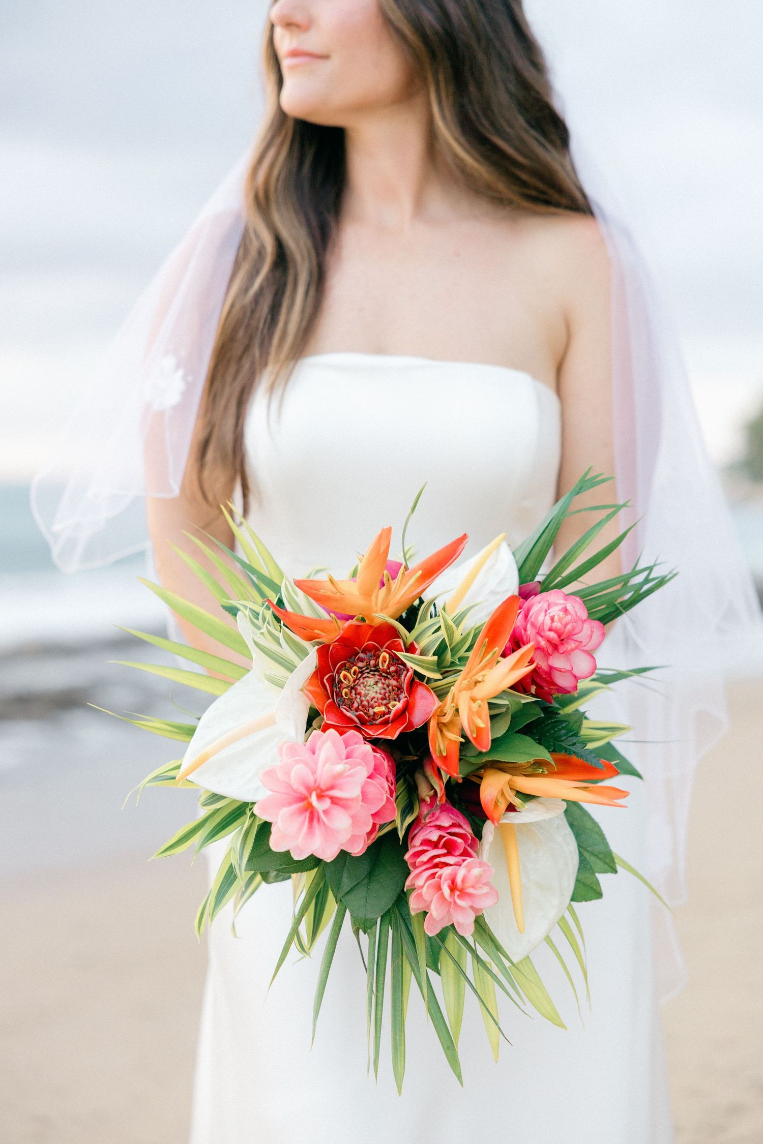 Tropical bridal bouquet featuring vibrant orange birds of paradise and pink flowers against a white dress.