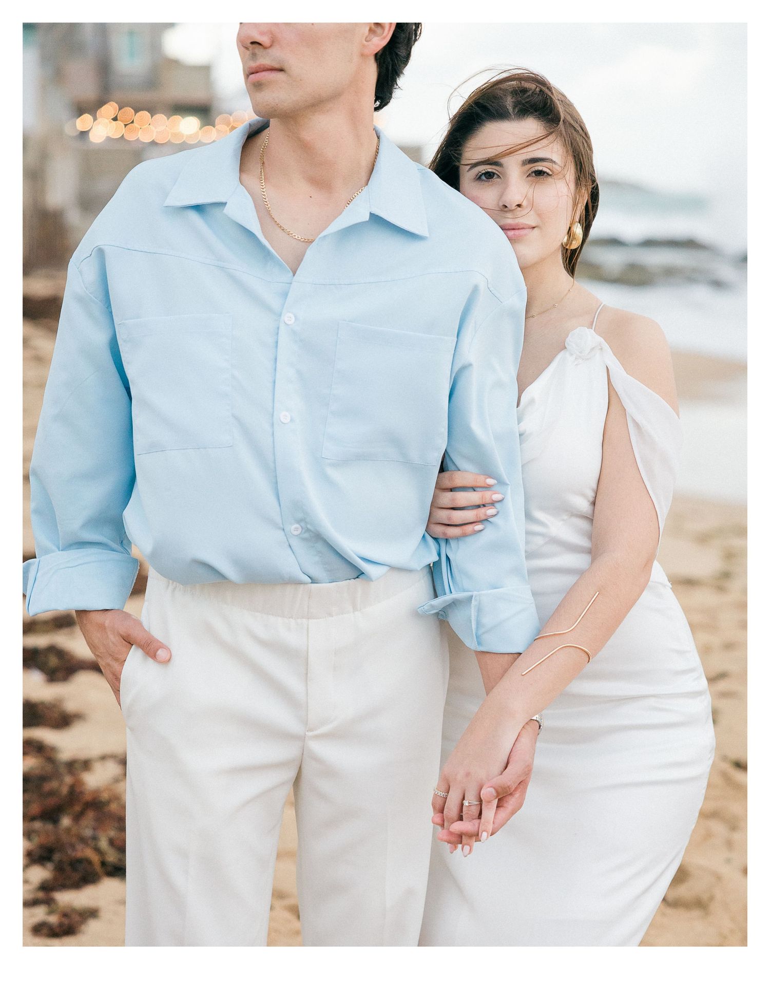A couple in light blue and white clothing poses together on a beach.