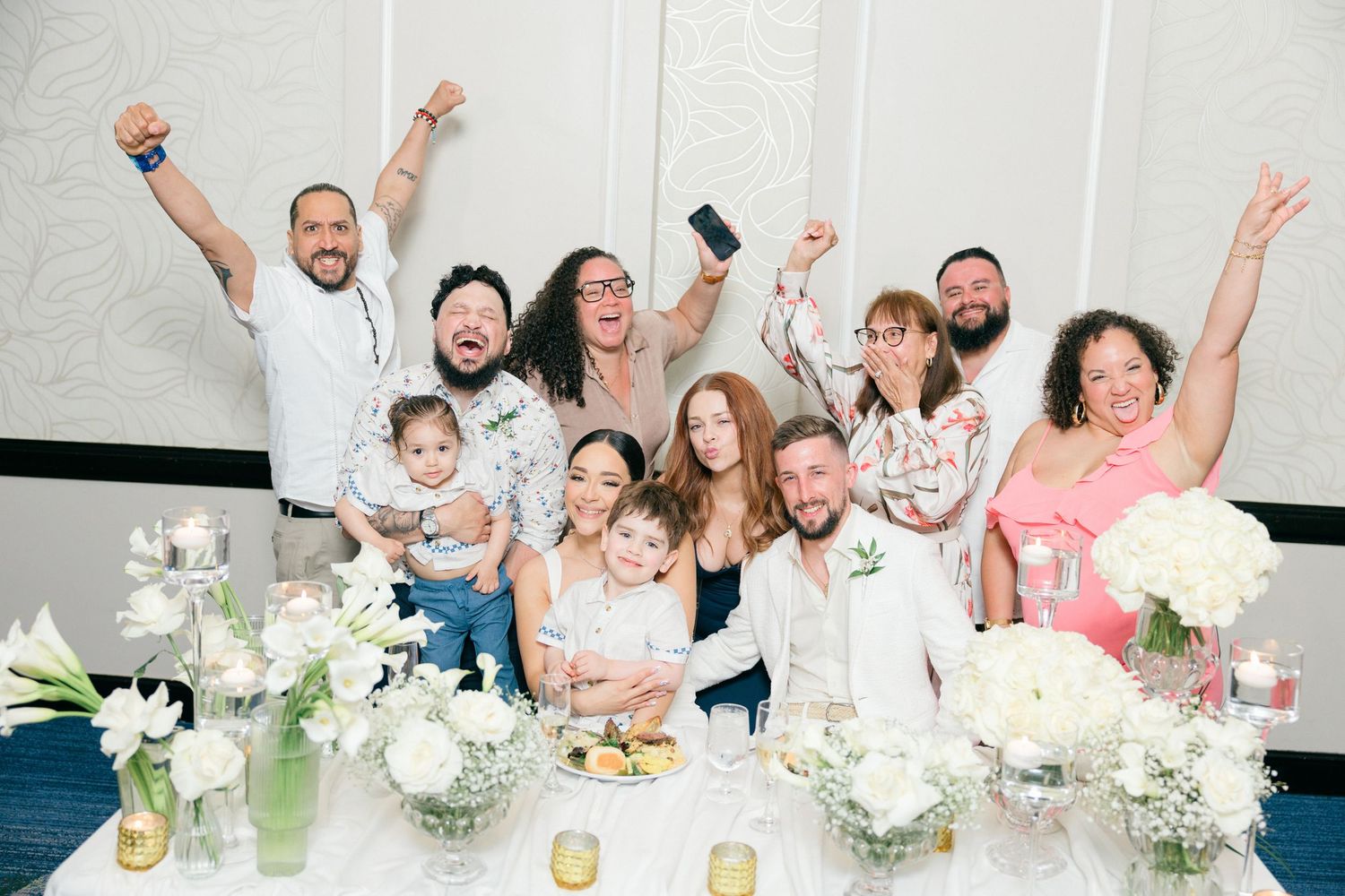 Celebratory group photo of wedding party raising hands in excitement at reception table.