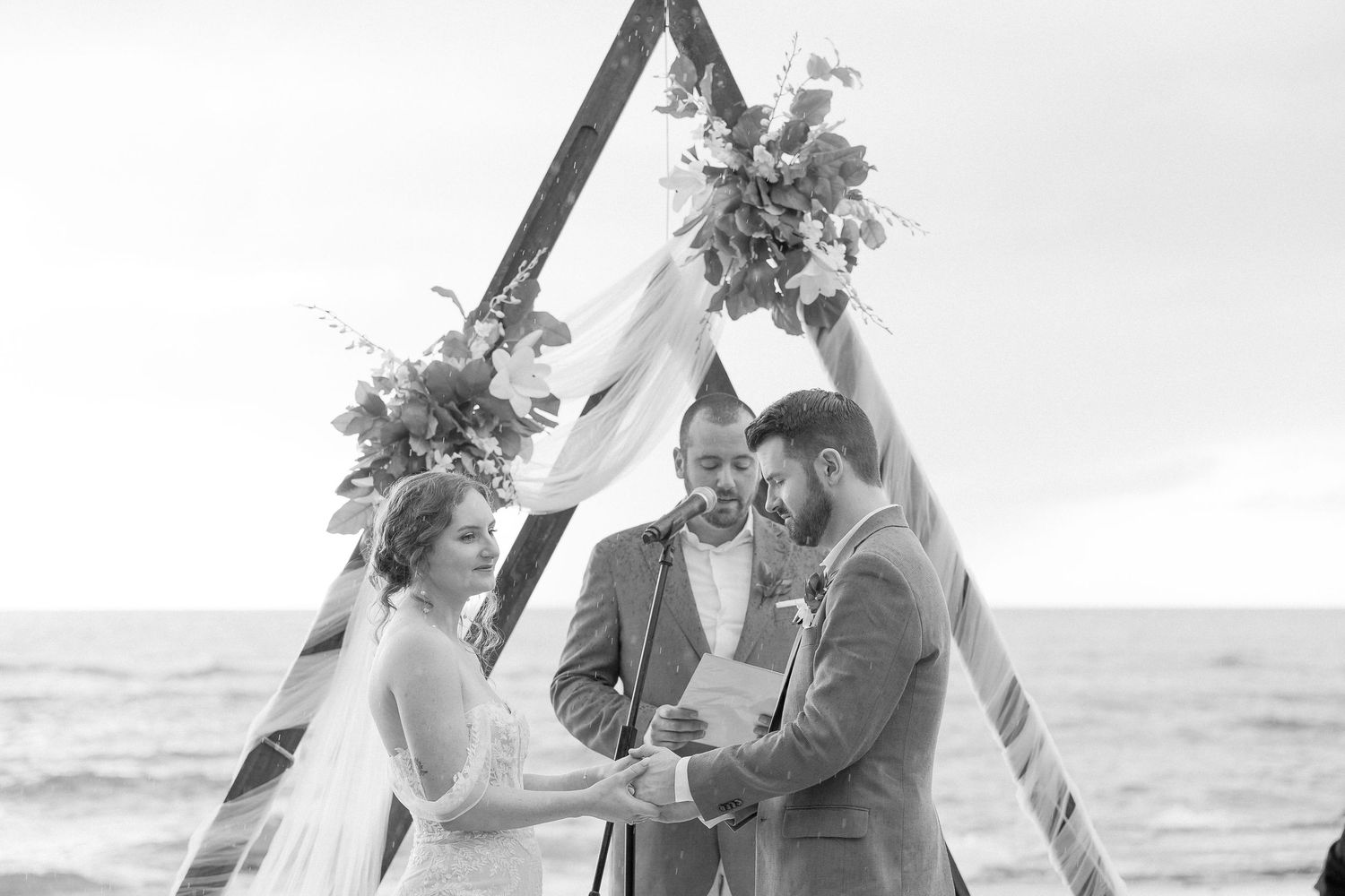 Black and white photo of a beach wedding ceremony under an arch with ocean views.