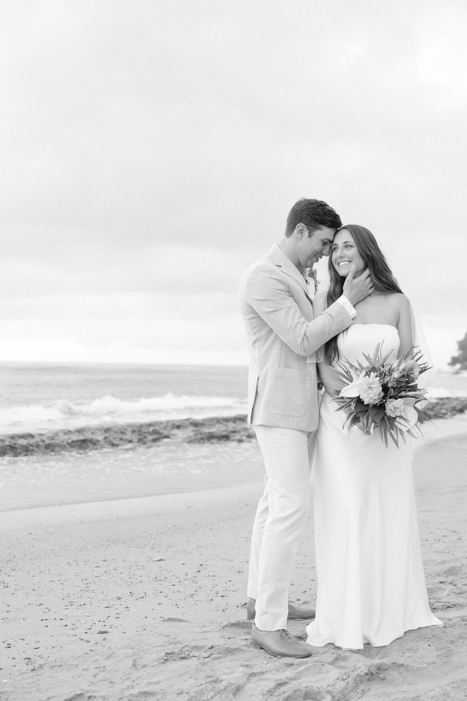 Black and white photo of couple sharing romantic moment on beach with tropical bouquet.