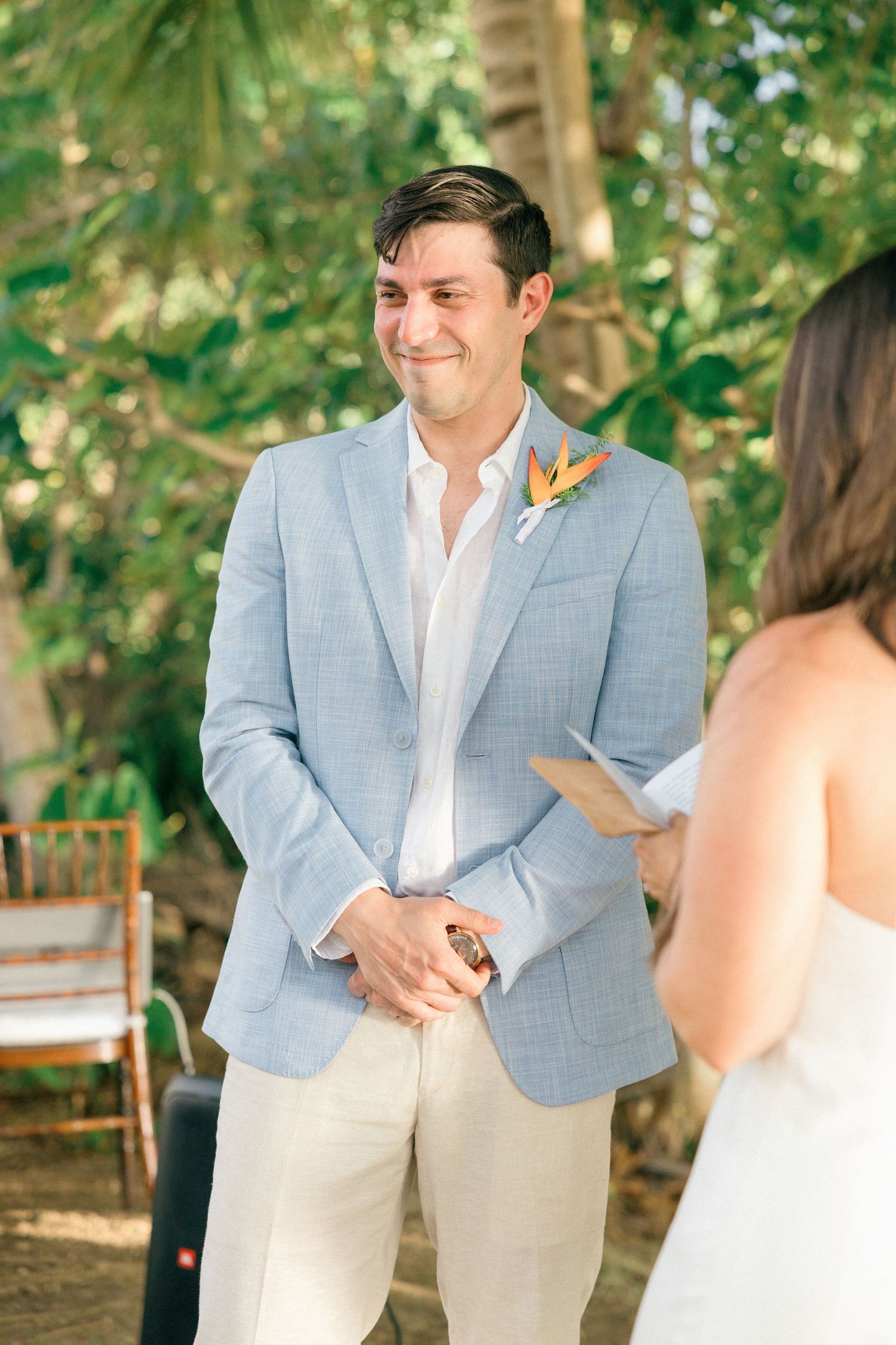 A groom in a light blue blazer and white pants reacts emotionally during an outdoor wedding ceremony.