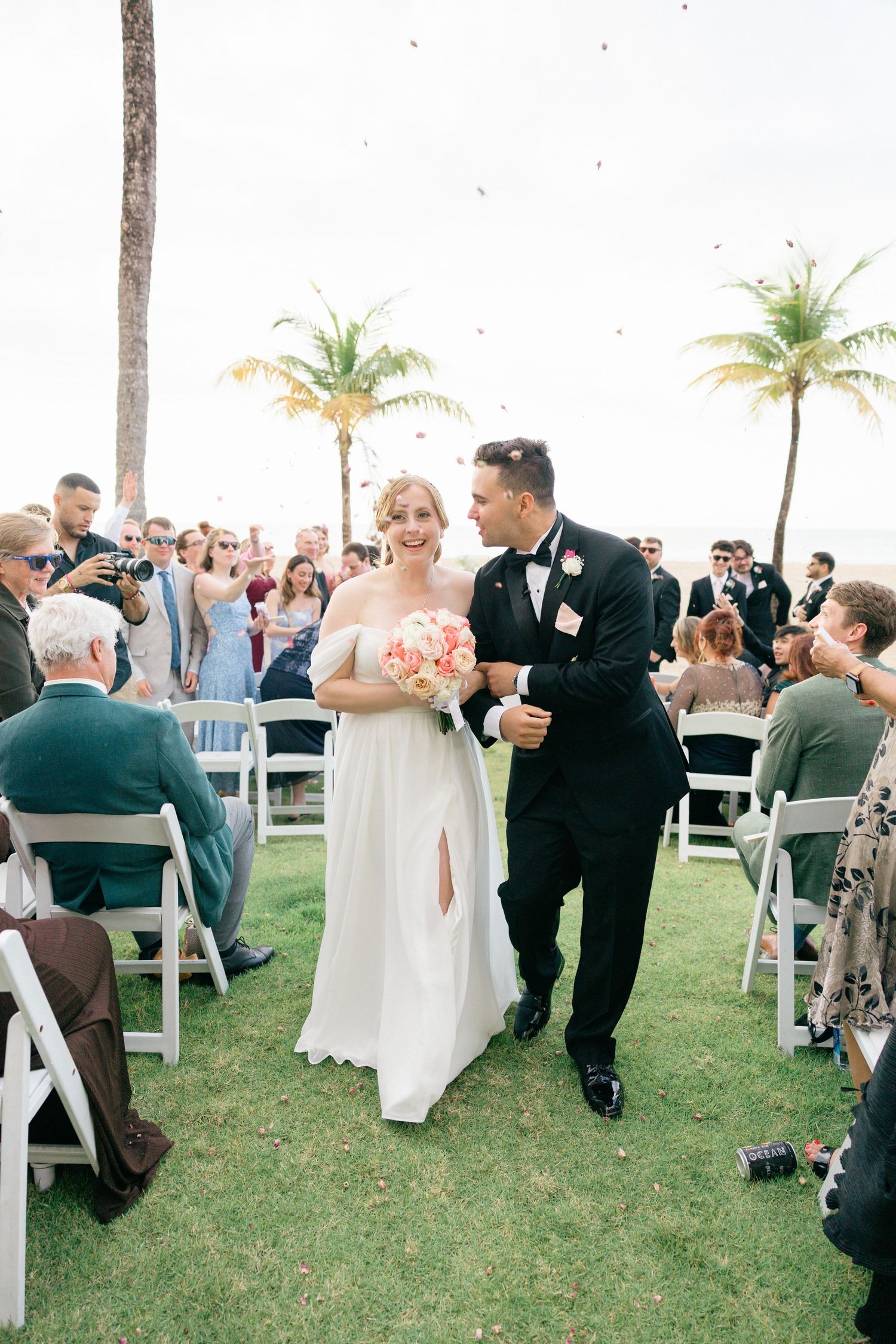 Wedding guests seated on white chairs enjoy an outdoor reception under palm trees.