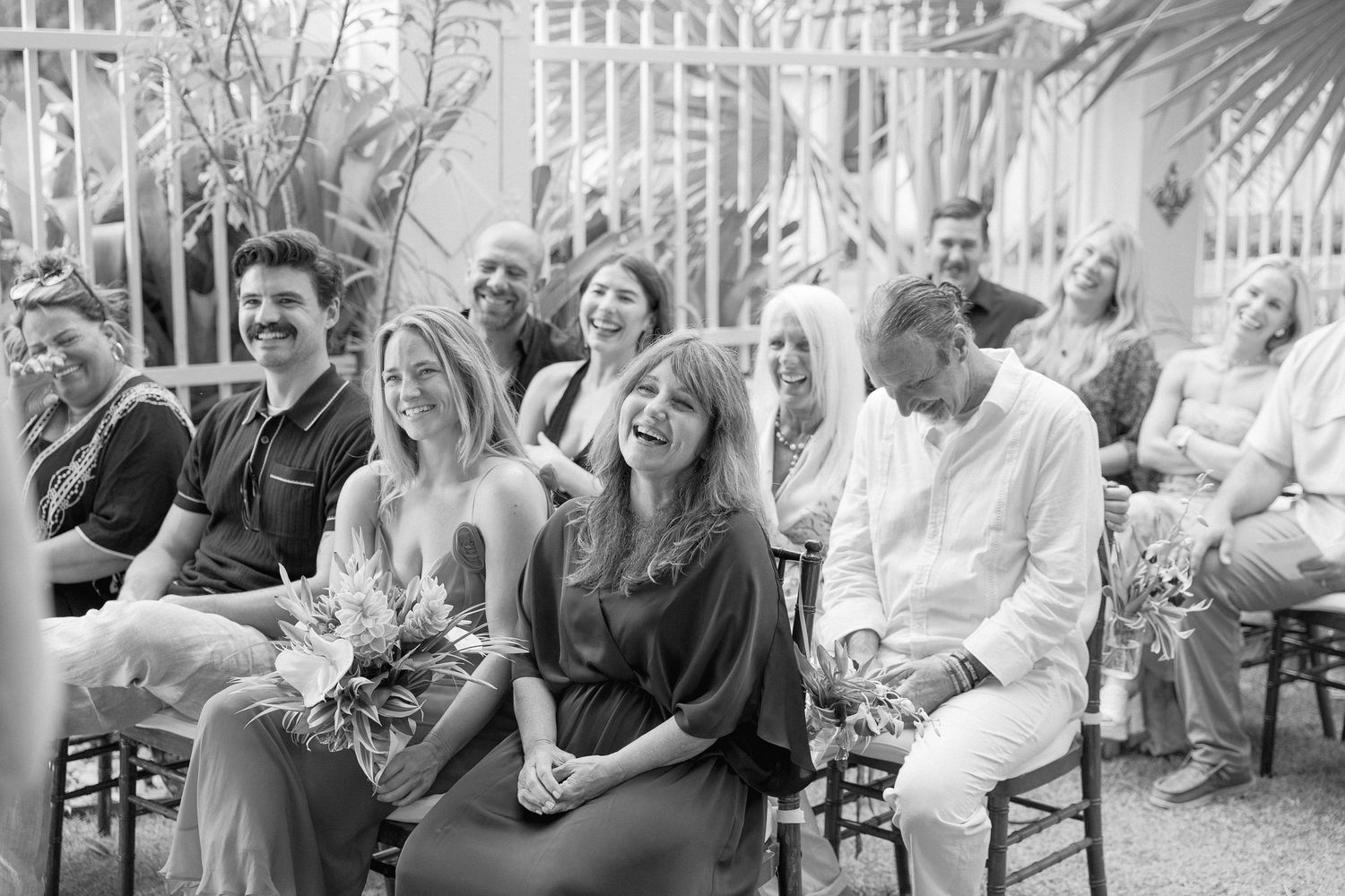 Wedding guests seated outdoors share joyful laughter during black and white ceremony moment.