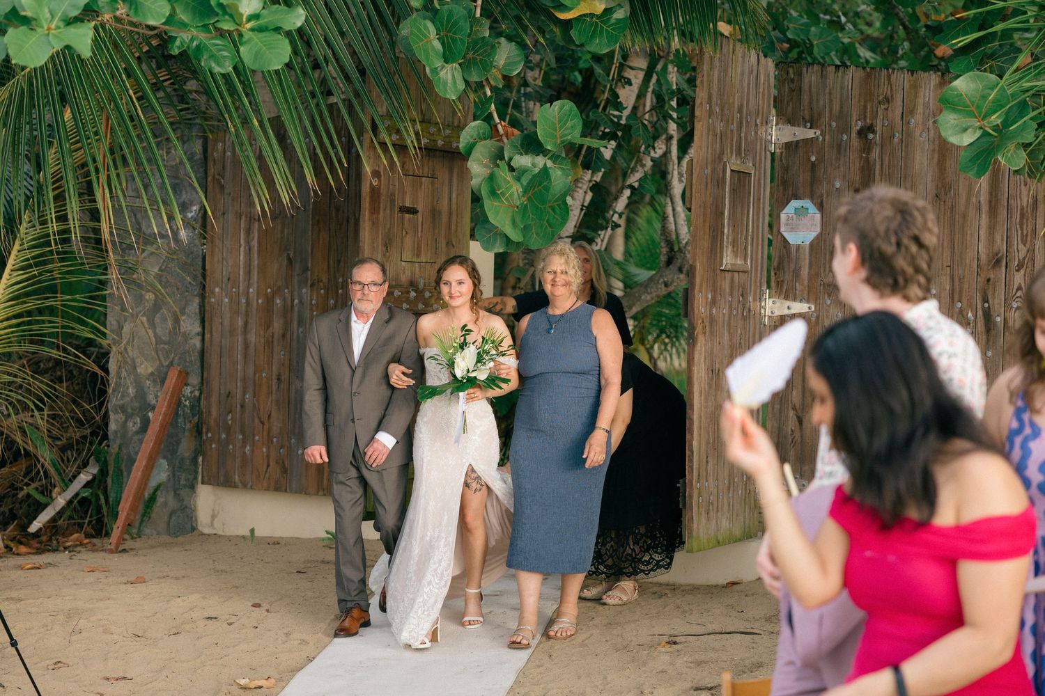 Wedding guests and party members gather on a tropical beach with wooden backdrop and greenery.