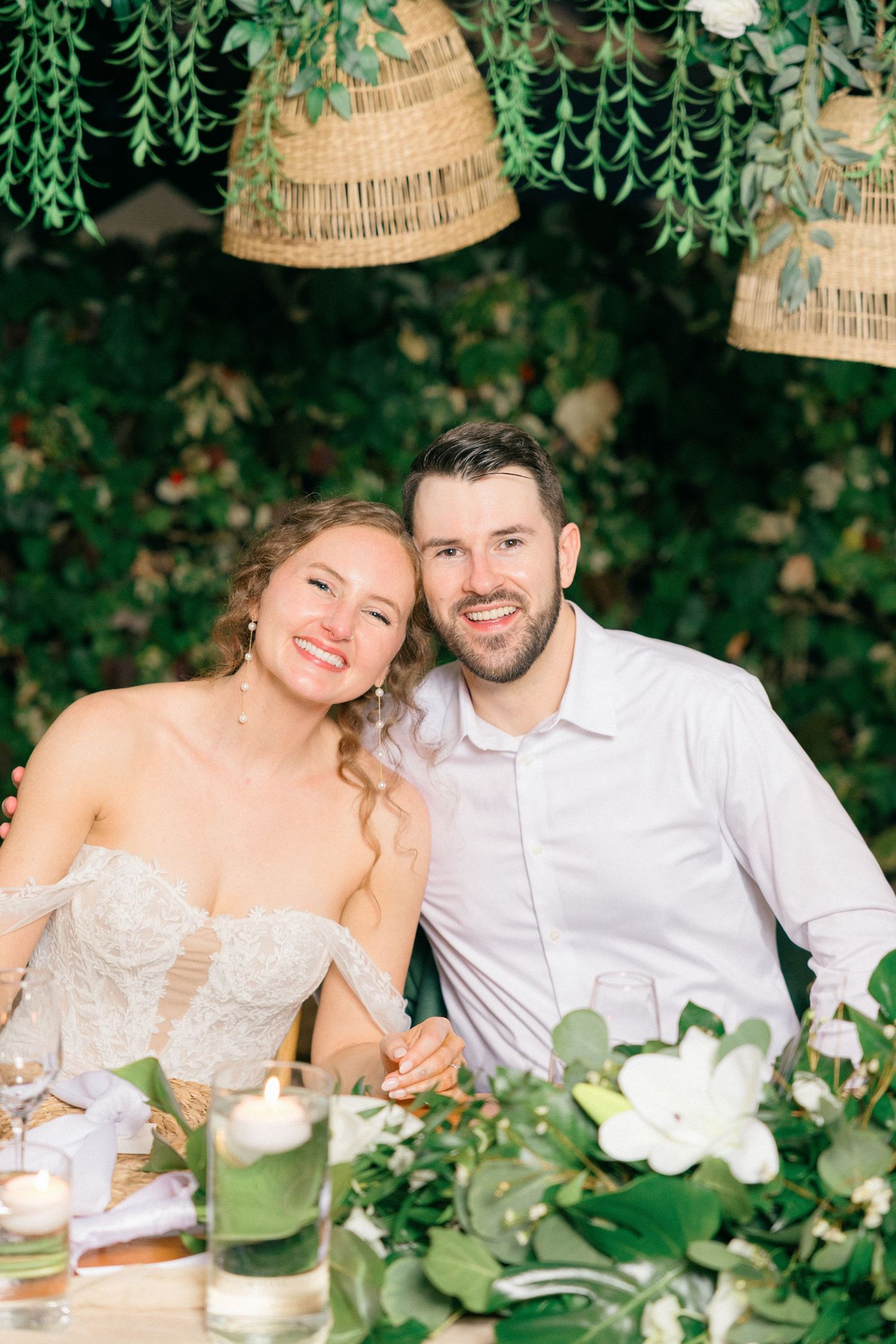 Couple sits at decorated outdoor reception table with hanging rattan lamps and greenery.