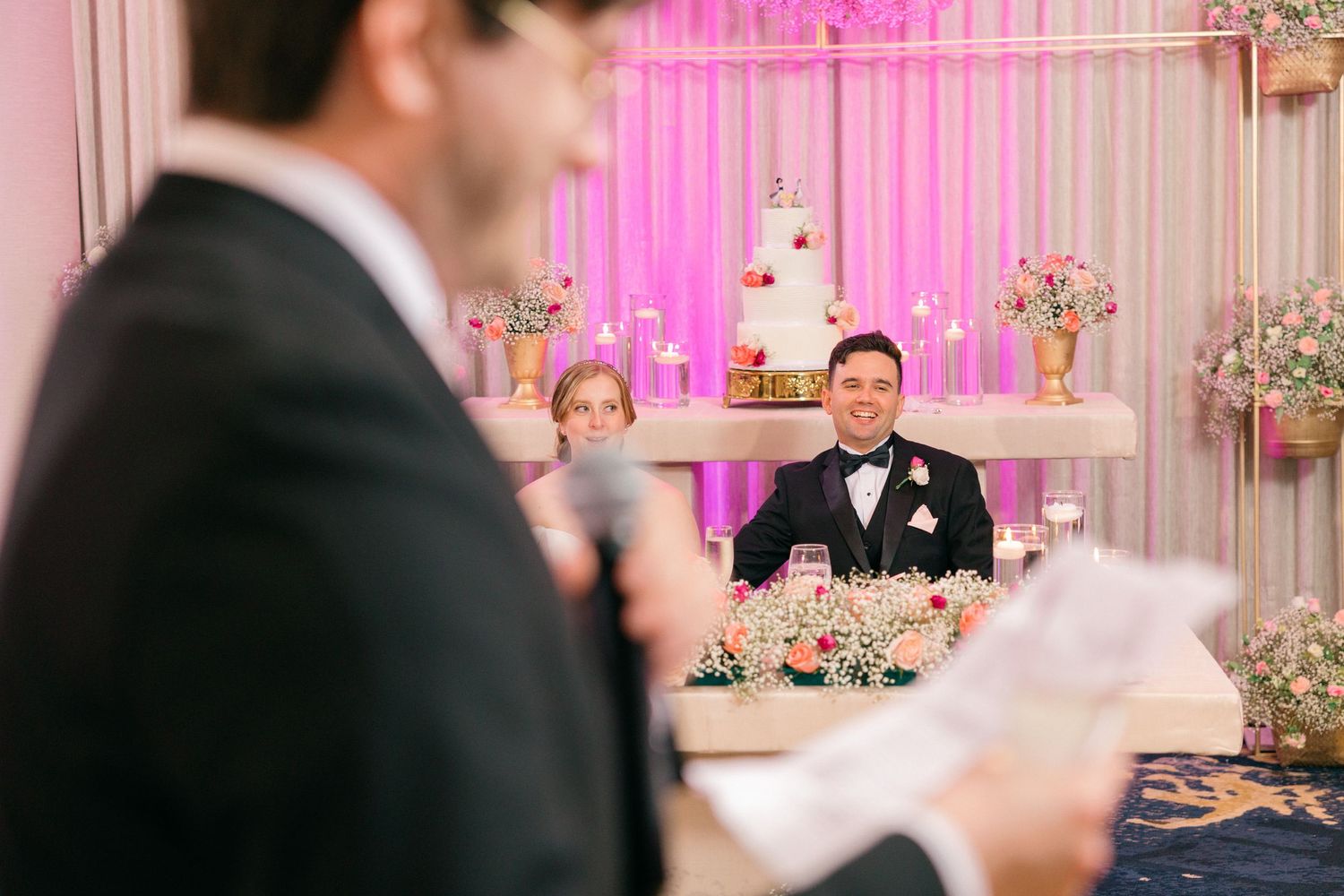 Wedding reception ballroom decorated with pink lighting and white drapery.