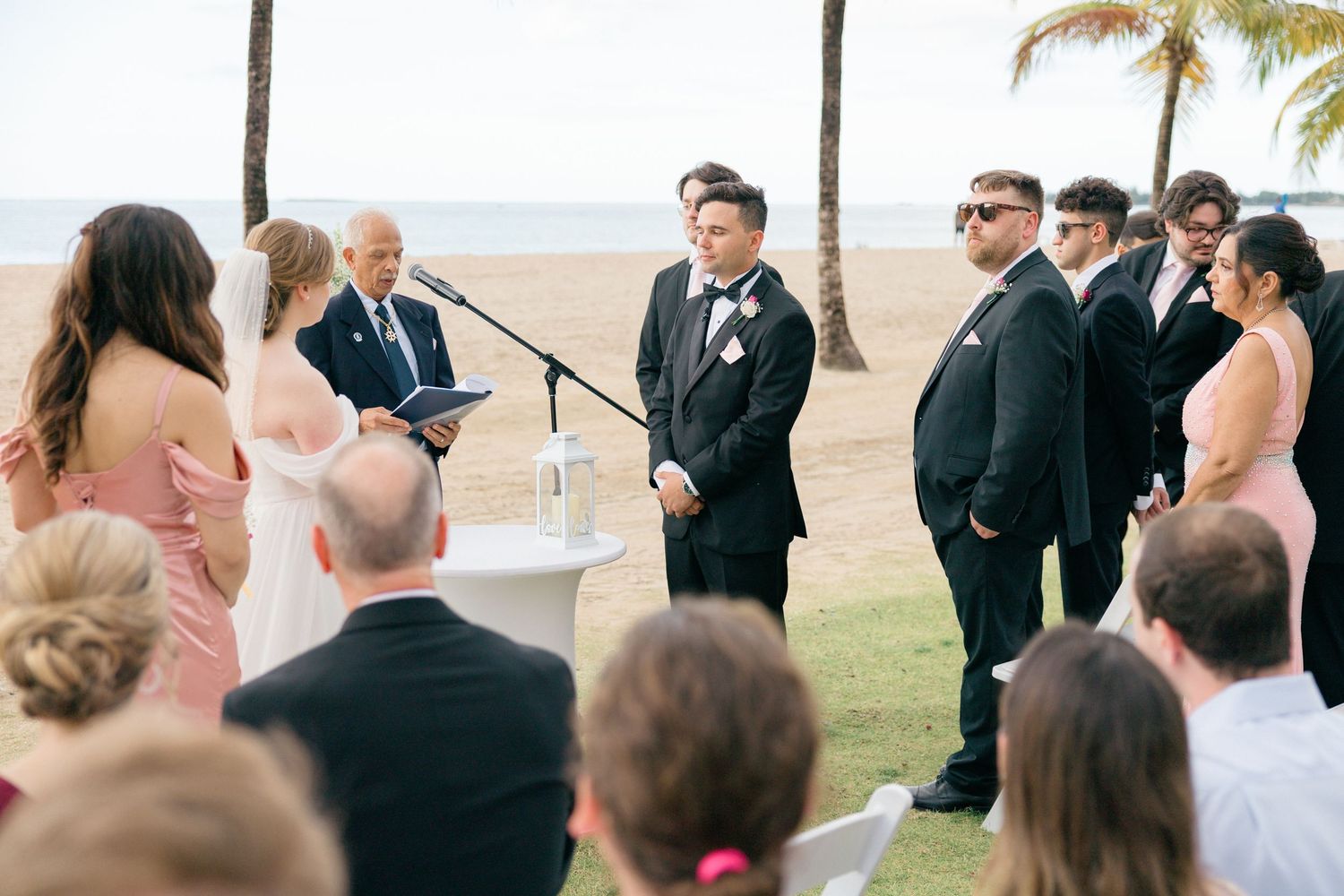 Beachside wedding ceremony with guests seated and palm trees swaying in background.