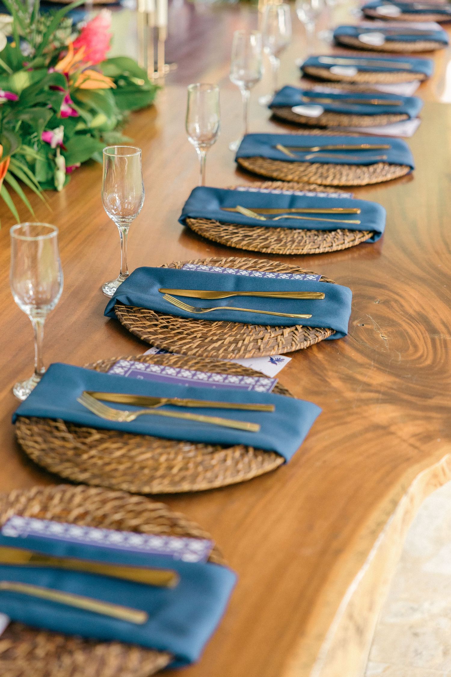 Elegant table setting with blue napkins, woven placemats, gold cutlery and champagne glasses on a wooden surface.