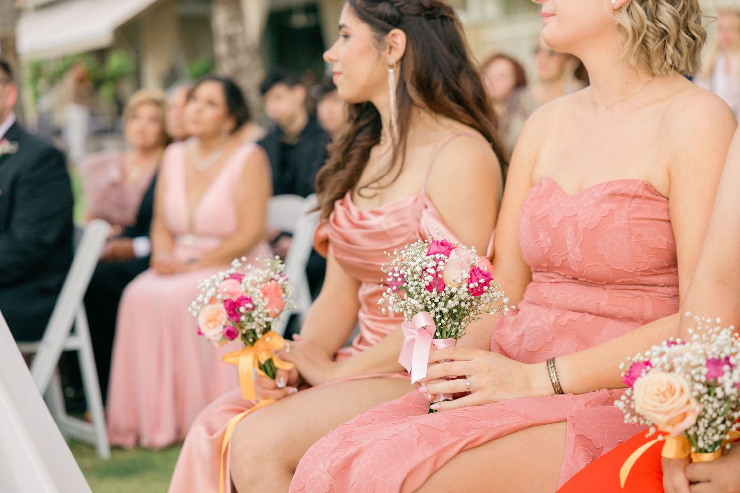 Bridesmaids in coral pink dresses hold delicate floral bouquets during an outdoor wedding ceremony.