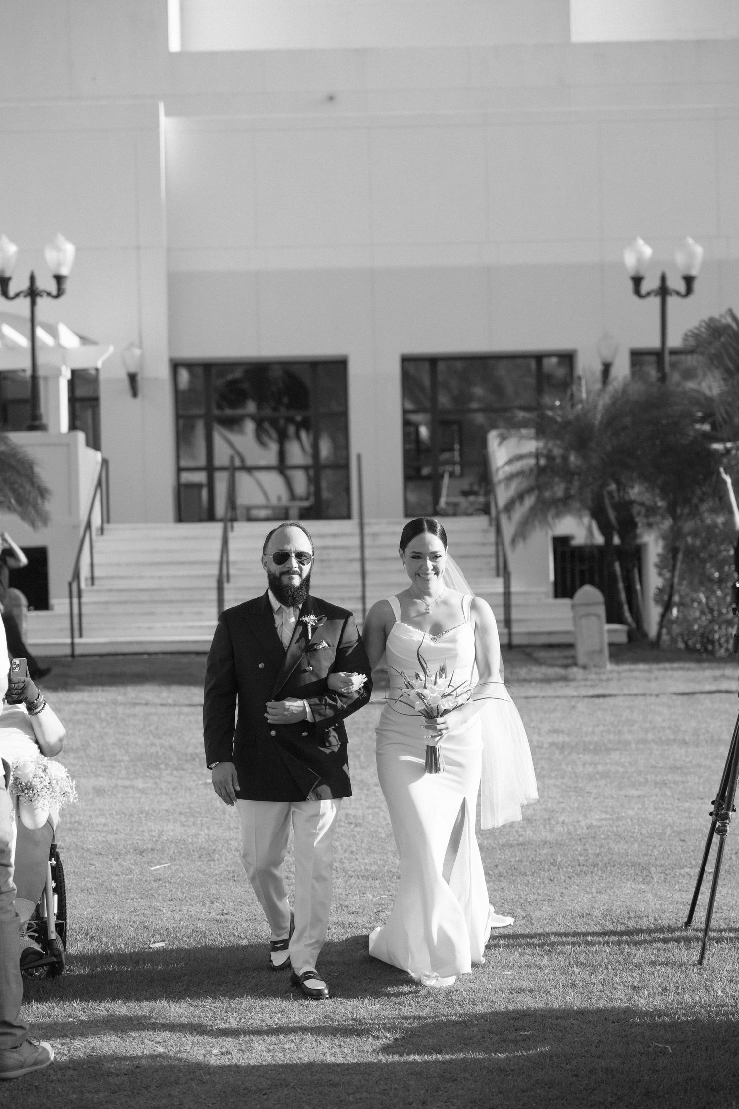Black and white photo of an outdoor wedding ceremony with guests seated on white chairs.