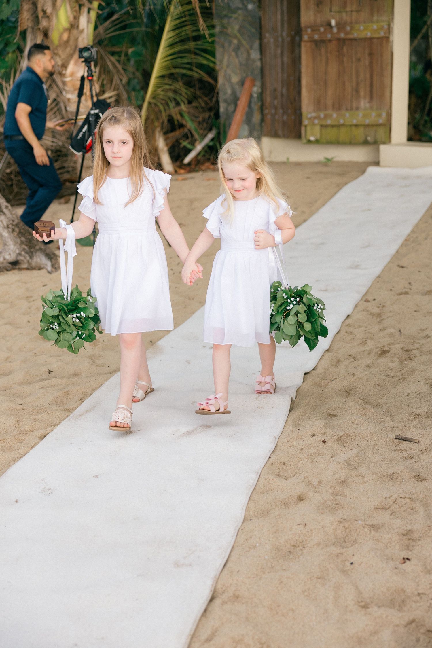 Two young flower girls in white dresses walk down a white aisle runner on a sandy beach.