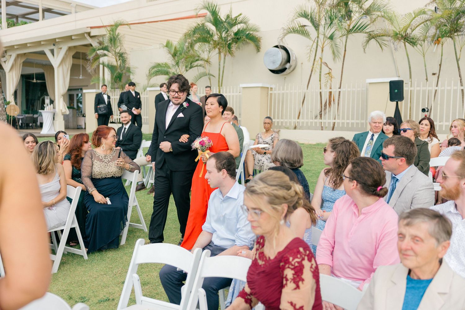Panoramic view of wedding guests seated outdoors with palm trees and white chairs.