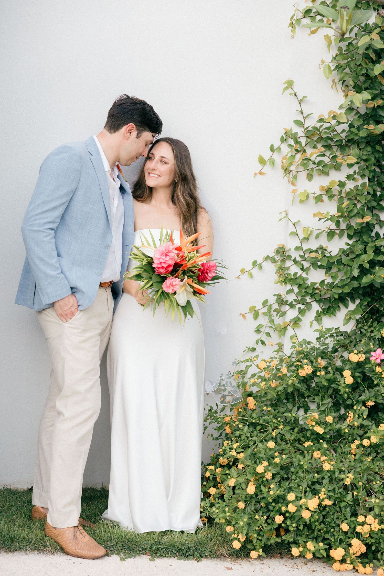 Wedding couple poses against white wall with yellow flowering vines.