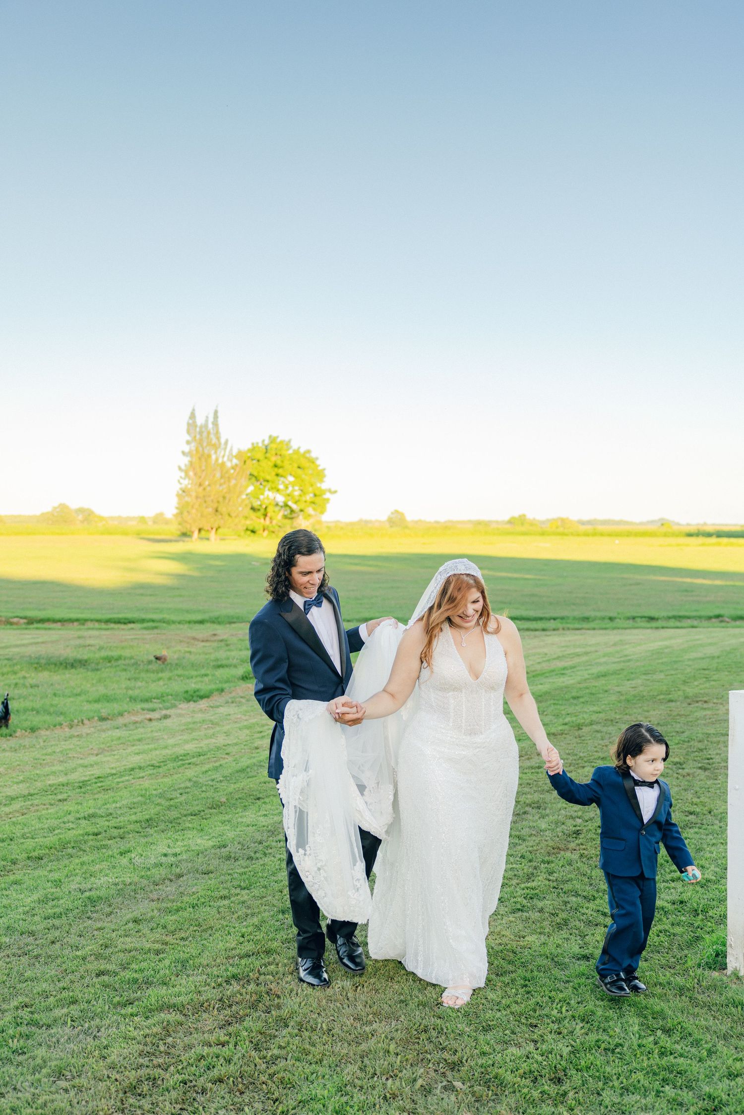 Wedding party walks together across a green field with flowing dress and small child.