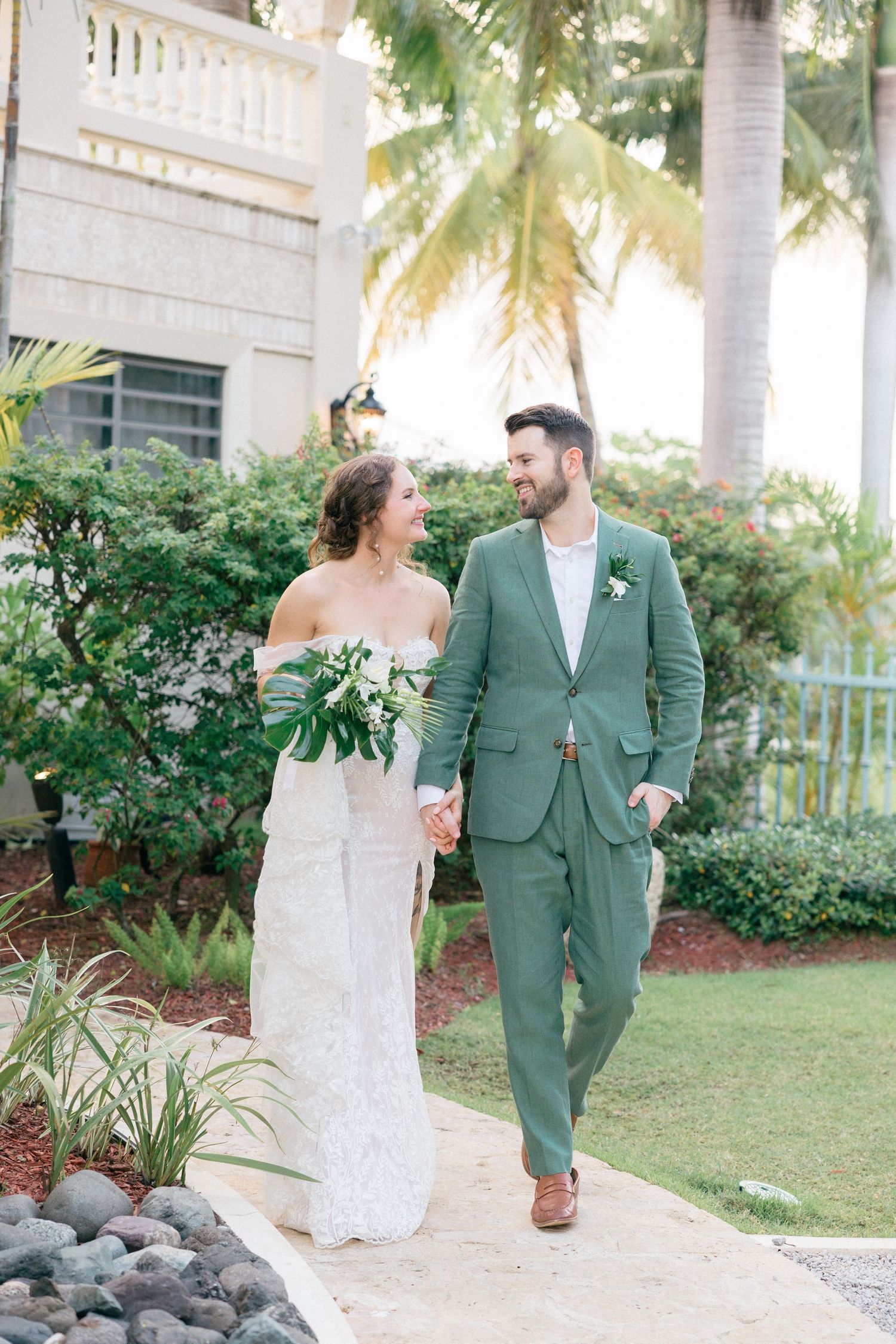 Couple in white dress and sage green suit walk together along a tropical garden path.
