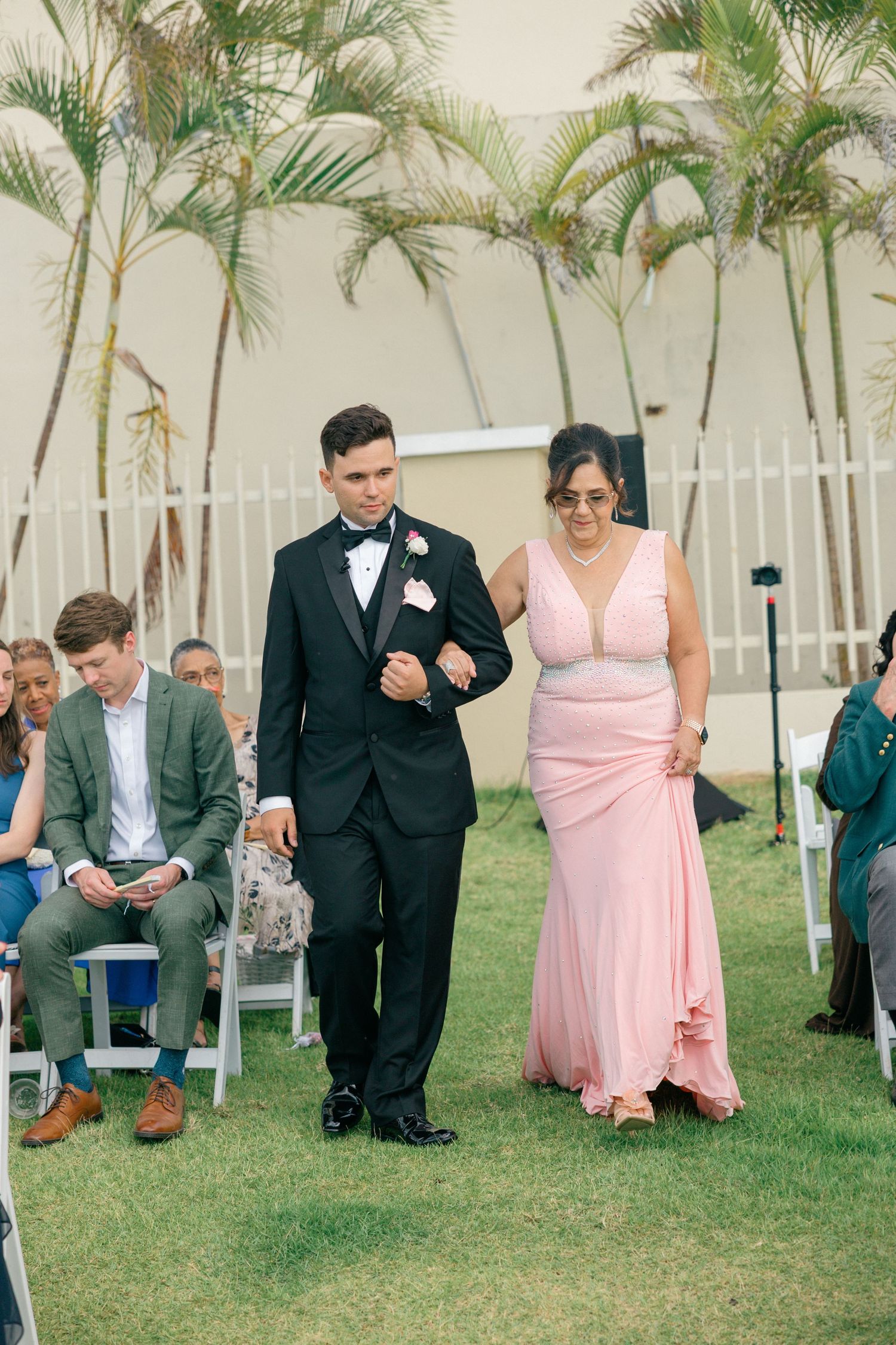 Wedding party members walking down an aisle lined with white chairs and palm trees.