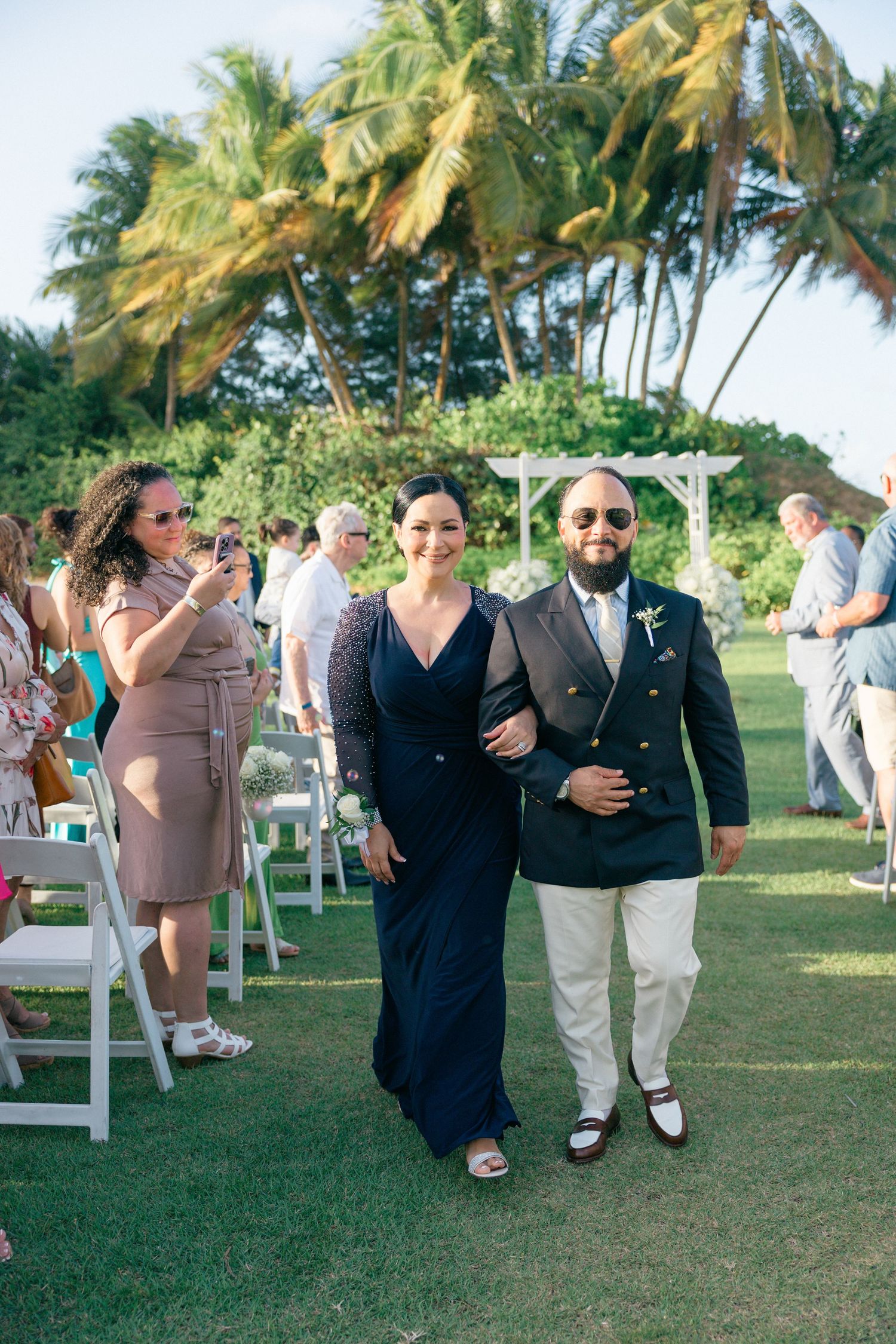 Wedding guests dance and celebrate on a tropical lawn with palm trees in the background.