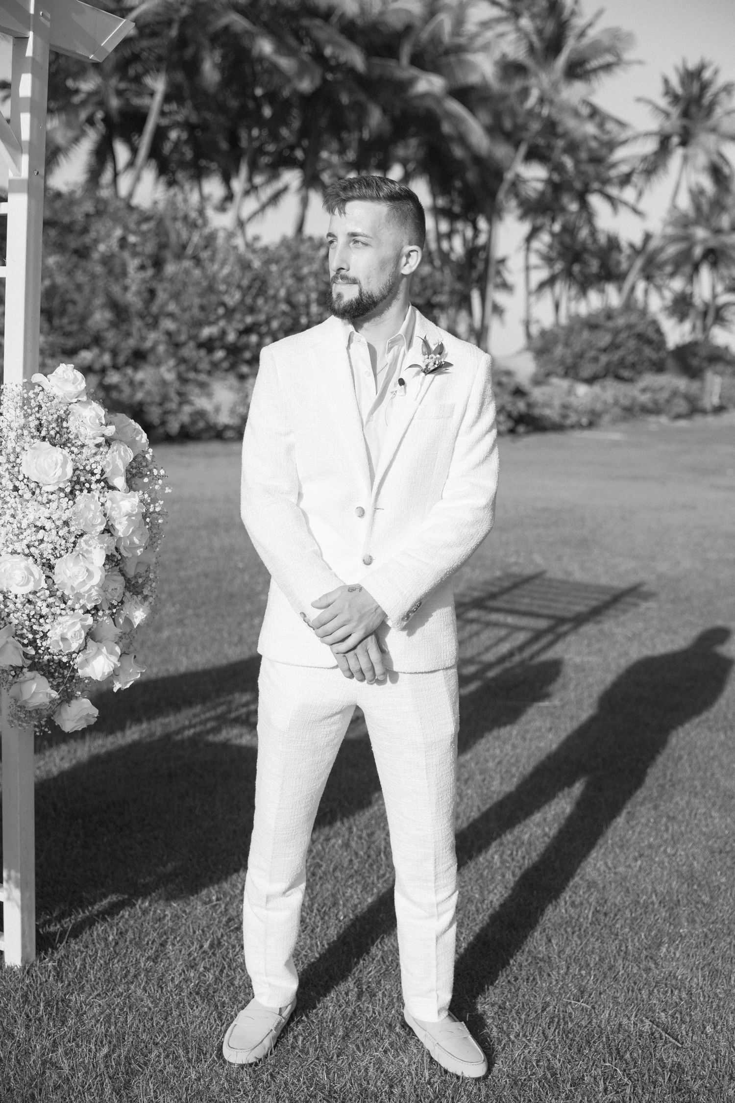 Wedding party members in white attire standing on grass with palm trees in background.