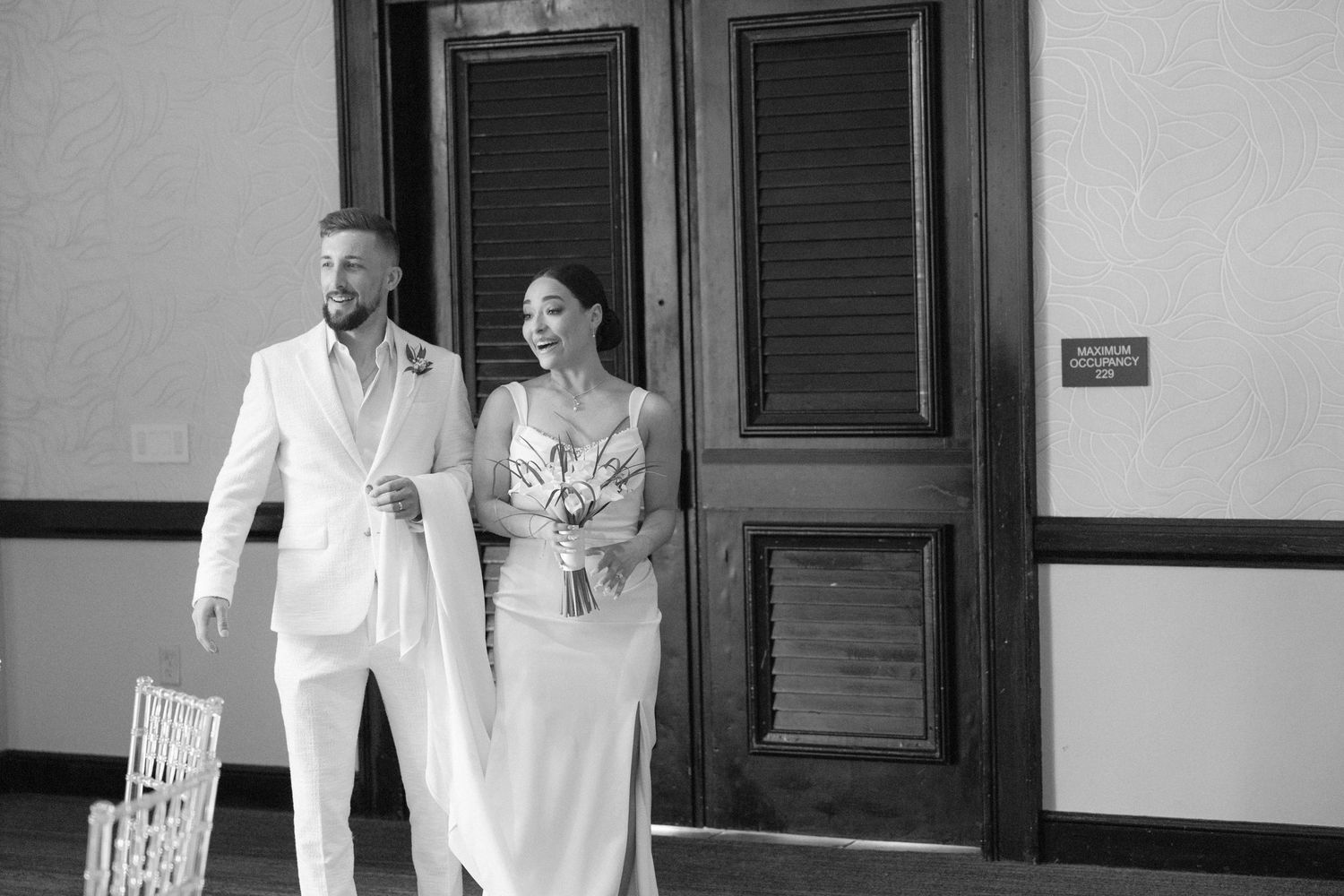 A couple in coordinated white outfits pose together in front of wooden doors.
