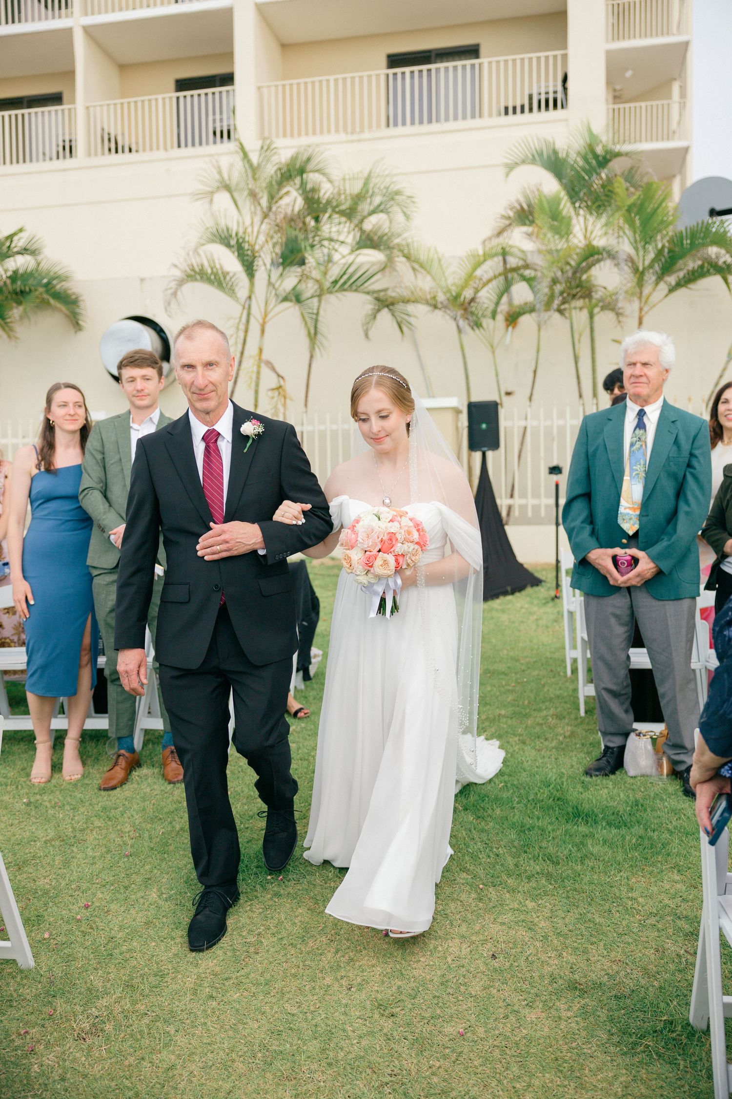 Wedding ceremony procession on a grassy lawn with a hotel building and palm trees in the background.