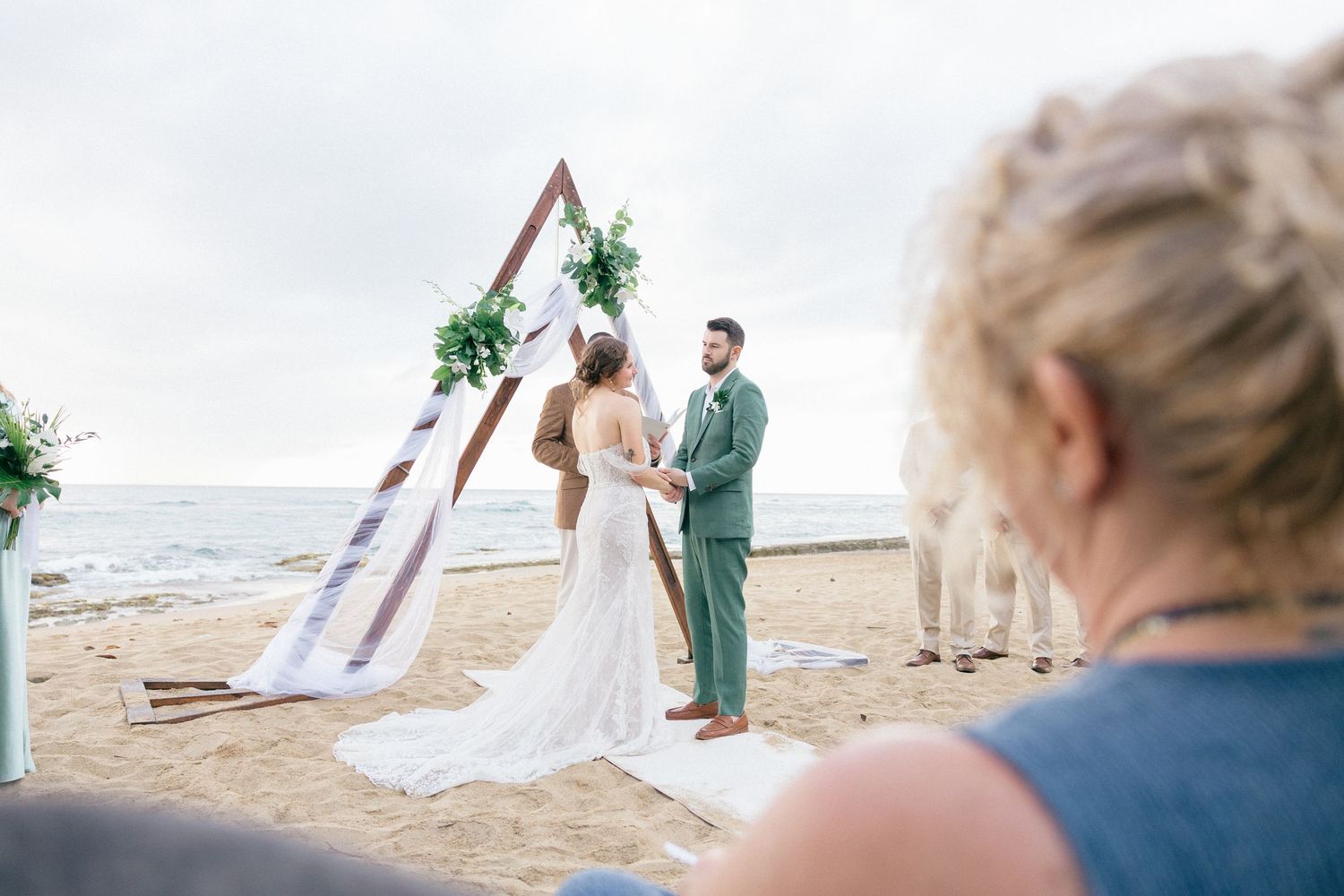 Romantic beach wedding ceremony with couple and guests gathered on sandy shore at sunset.