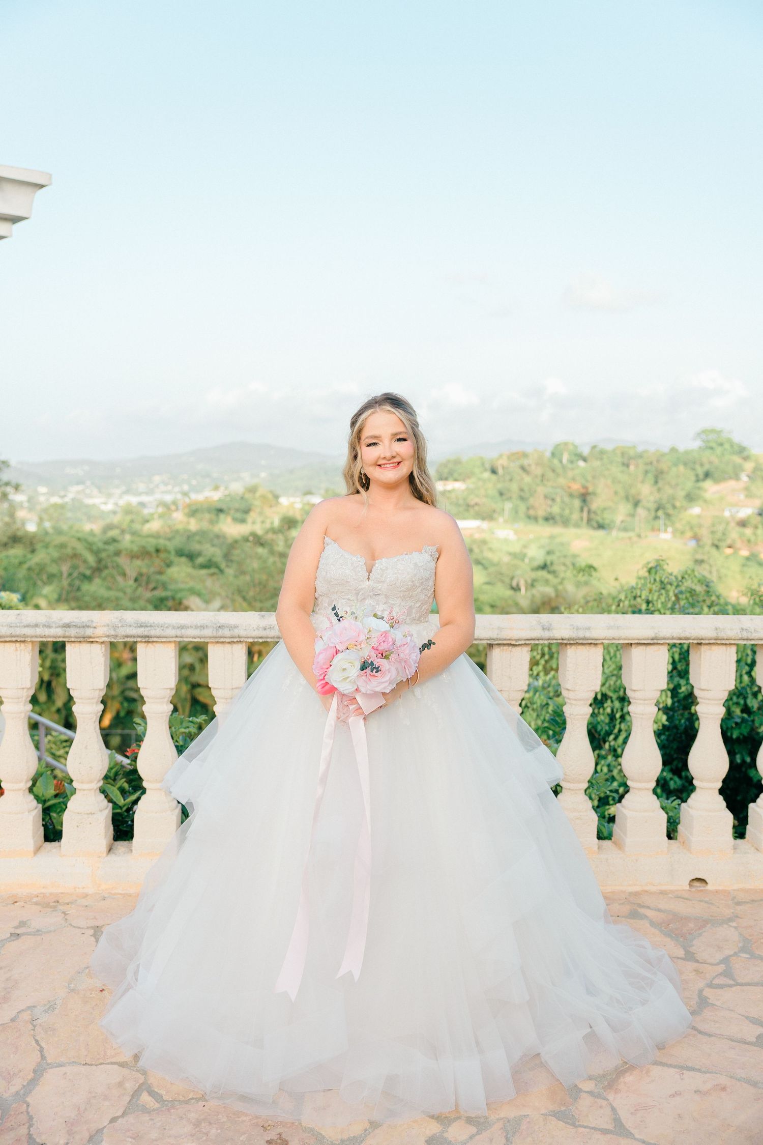 A flowing white wedding gown with tulle skirt against a stone balcony overlooking a scenic landscape.
