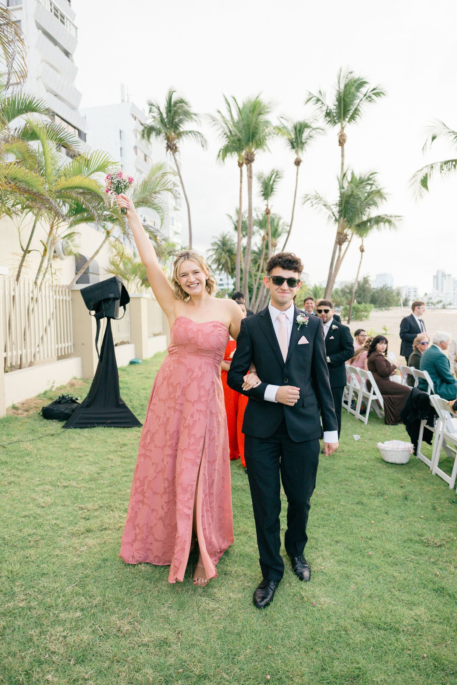 Wedding attendees in formal wear walk across grass during outdoor celebration.