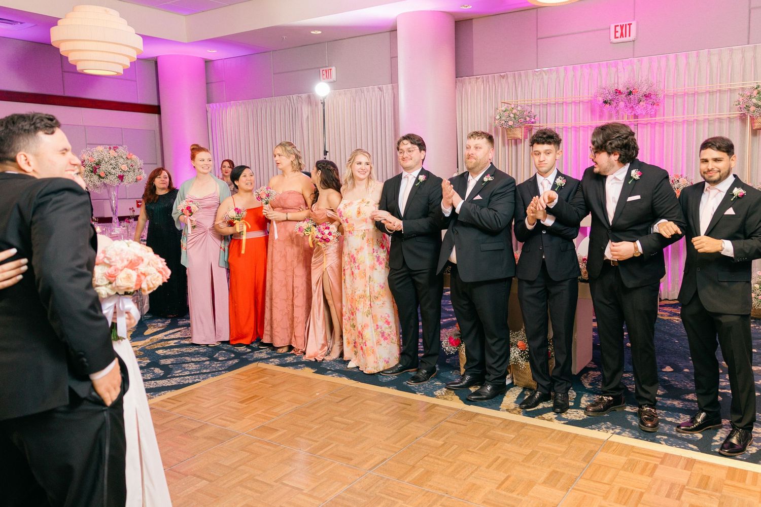 Wedding party members line up in formal attire at an elegant reception with pink lighting.
