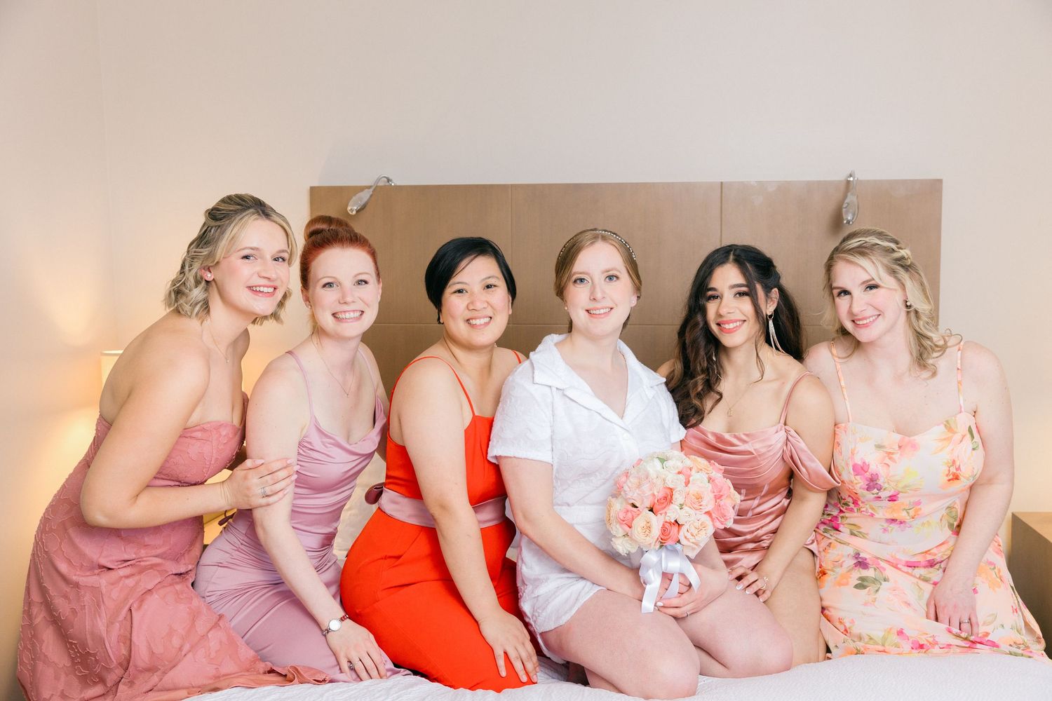 Wedding party wearing coordinated pink and coral dresses posing together on a bed.