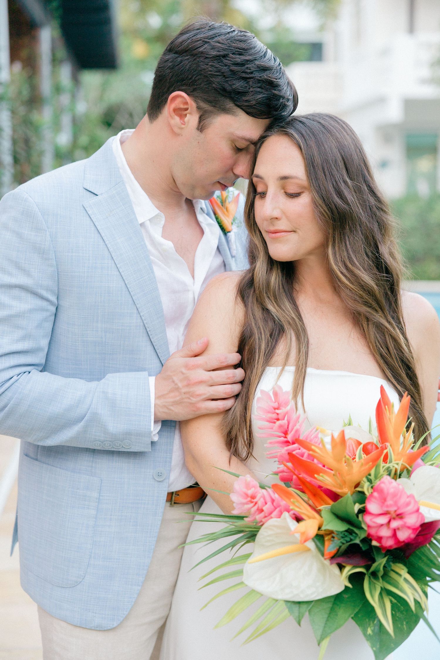 A romantic tropical wedding couple embraces while holding a vibrant bouquet with orange and pink tropical flowers.