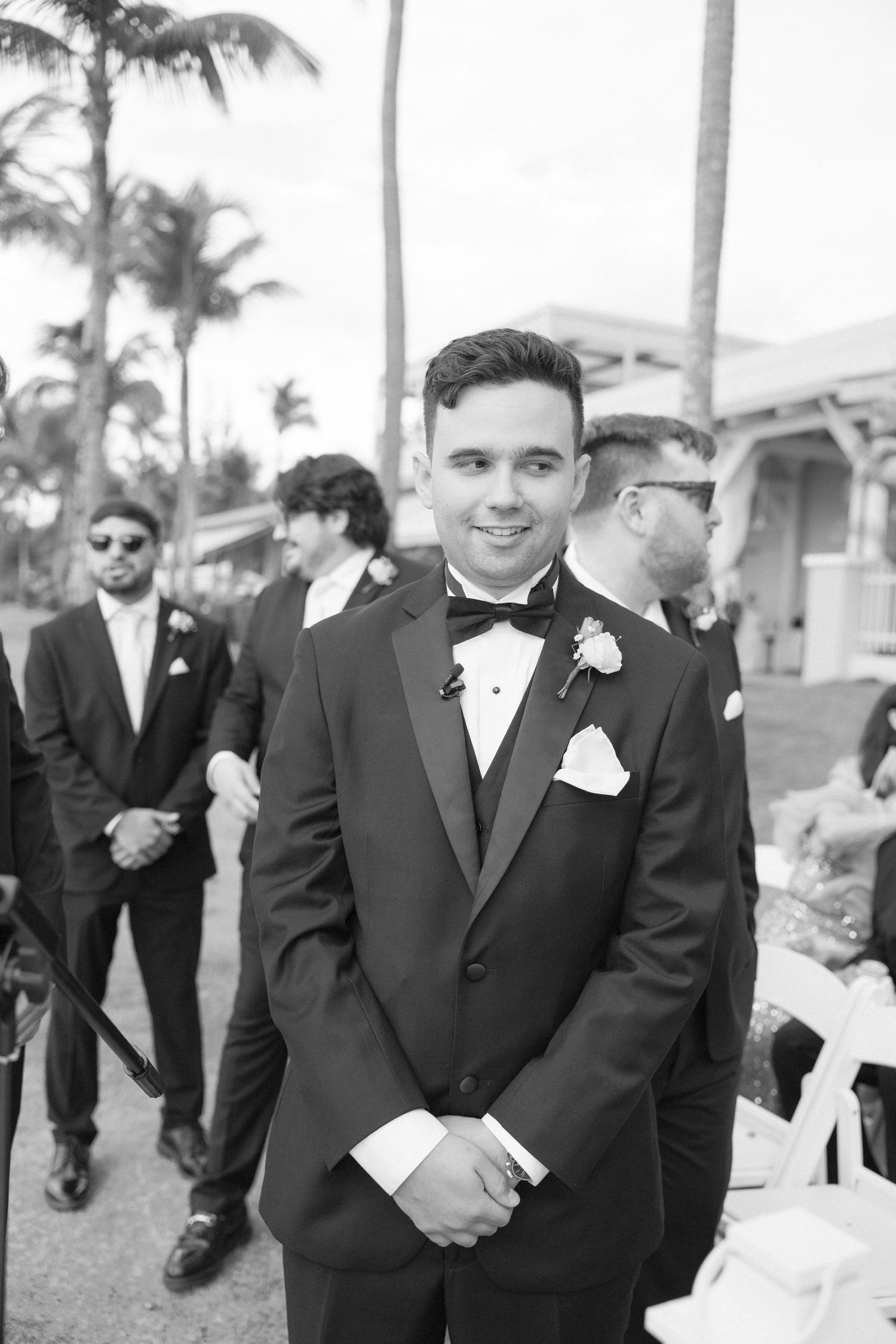 Wedding party members in formal black tuxedos stand outdoors with palm trees in background.