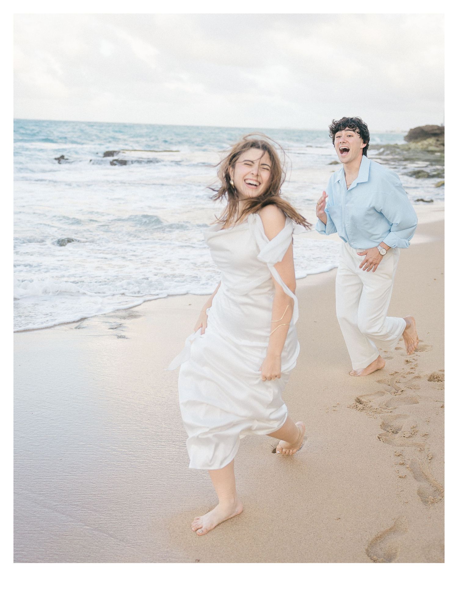 Joyful couple running and laughing on beach in casual wedding attire.