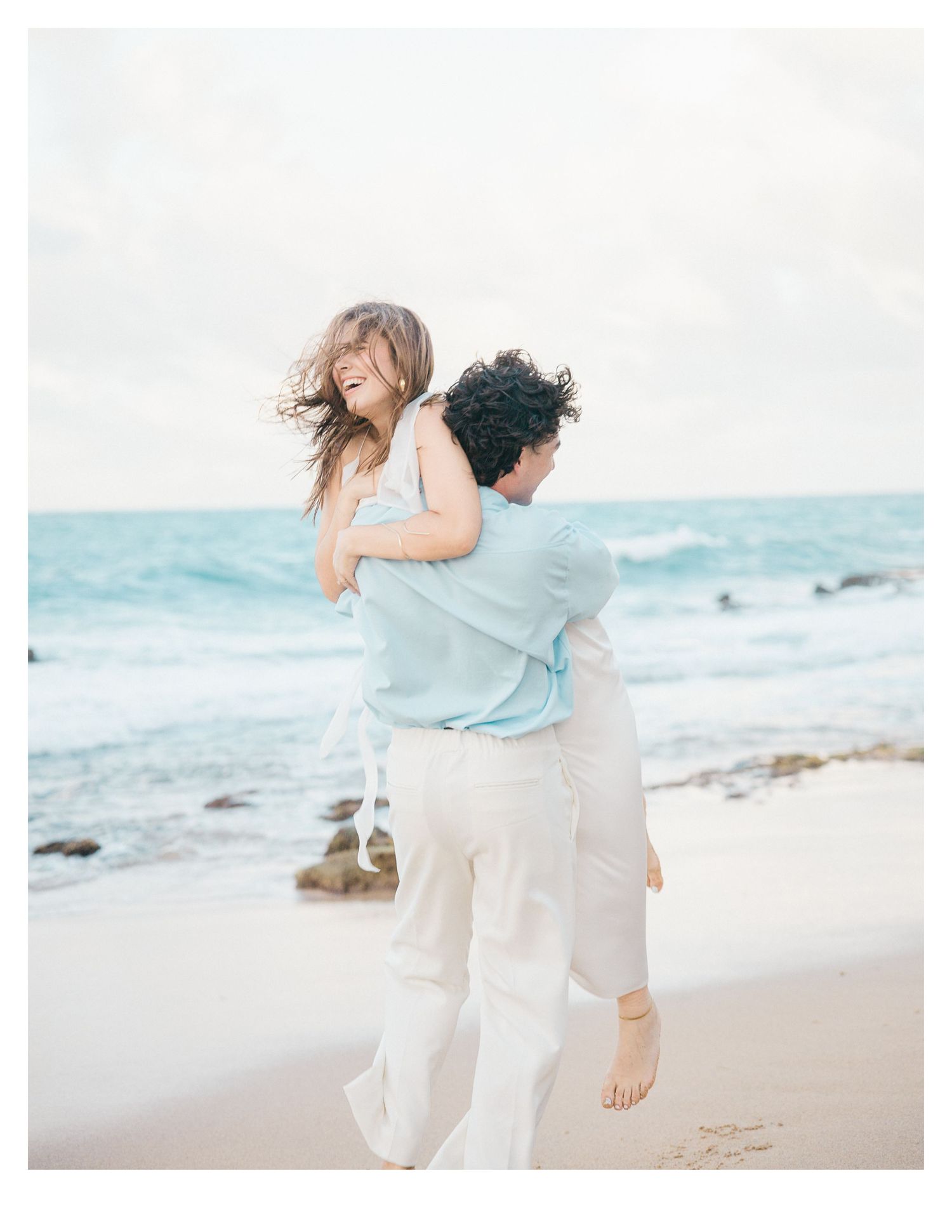 A couple in light colored clothing embraces on a beach with turquoise ocean waters.