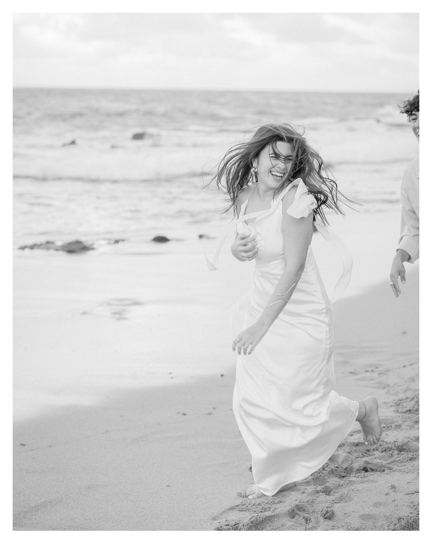 Woman in flowing white dress laughing with wind-swept hair on beach.