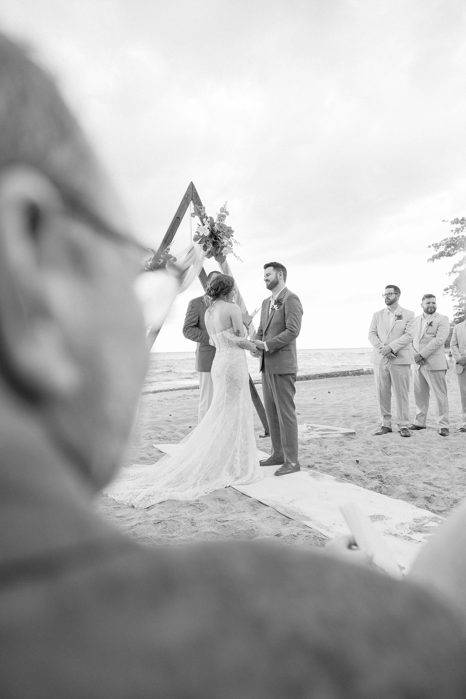 Black and white photo of beach wedding ceremony with guests watching couple exchange vows.