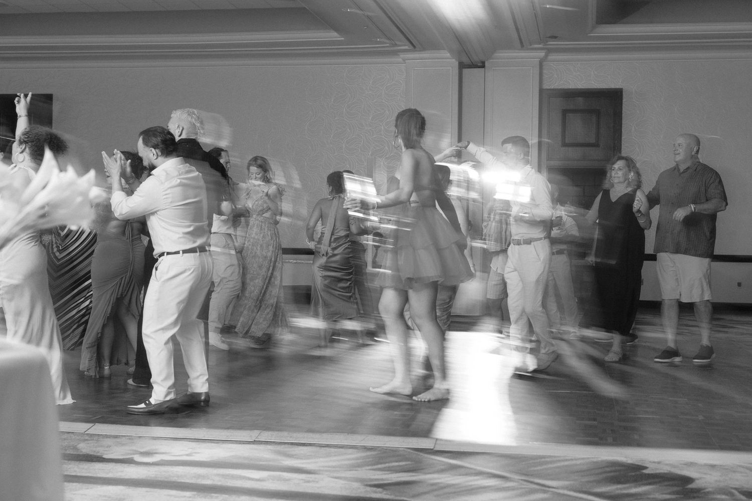 Wedding party dancing in a large reception hall with overhead lighting.