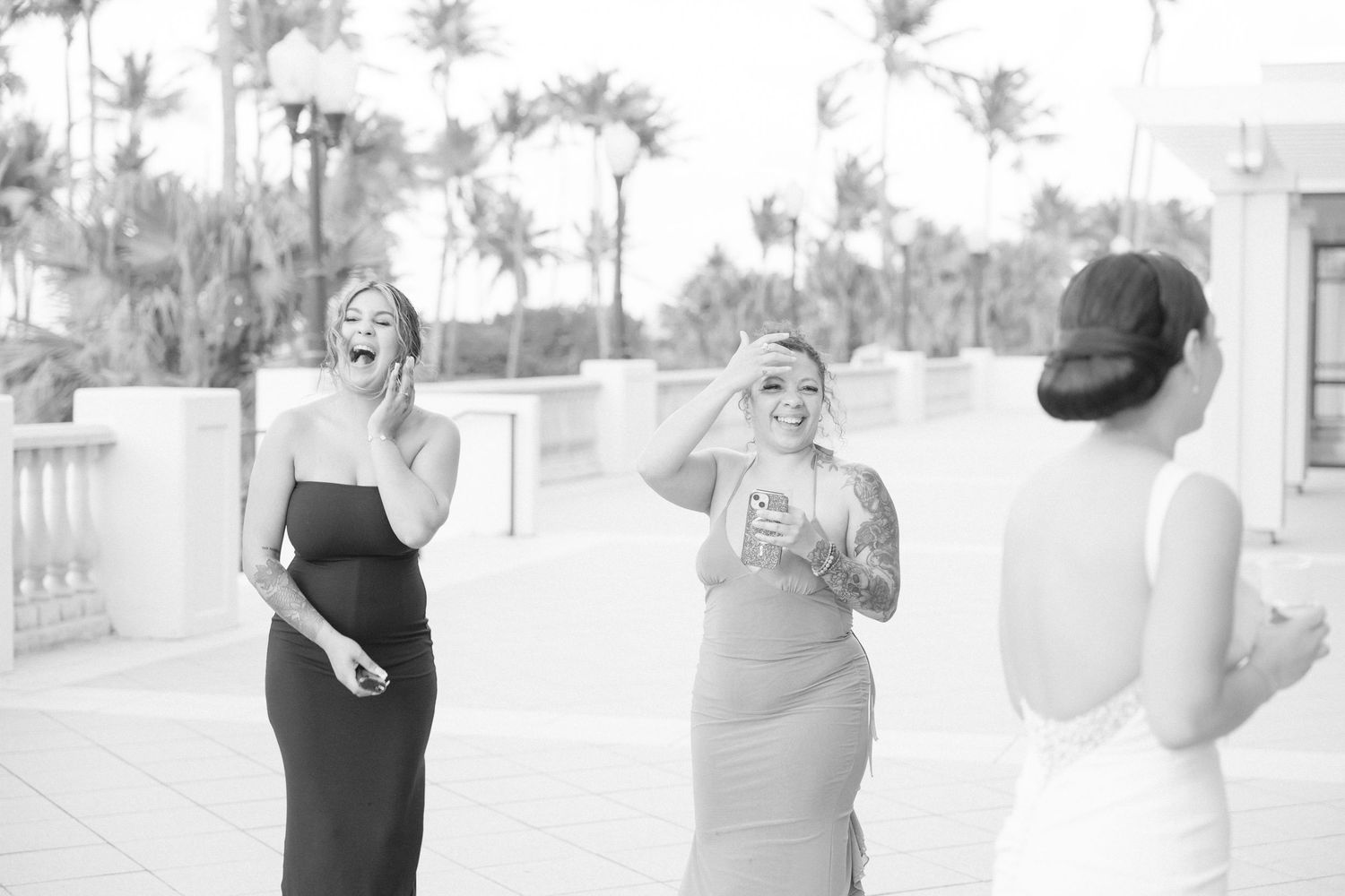 Three people in formal attire enjoying conversation on an outdoor terrace with palm trees.