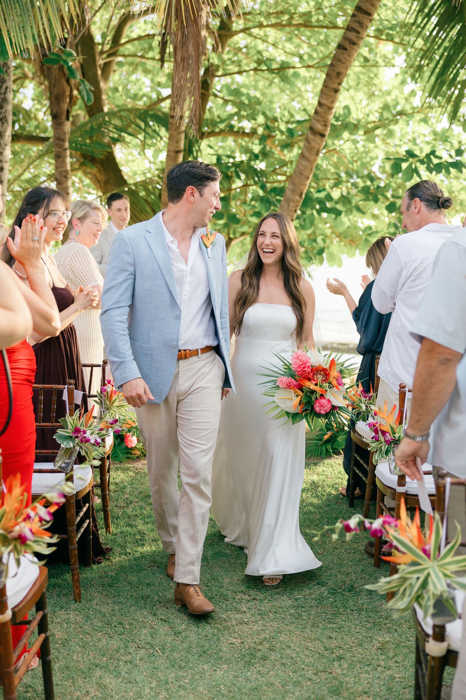 Tropical wedding ceremony setup with guests seated under palm trees and colorful floral arrangements.