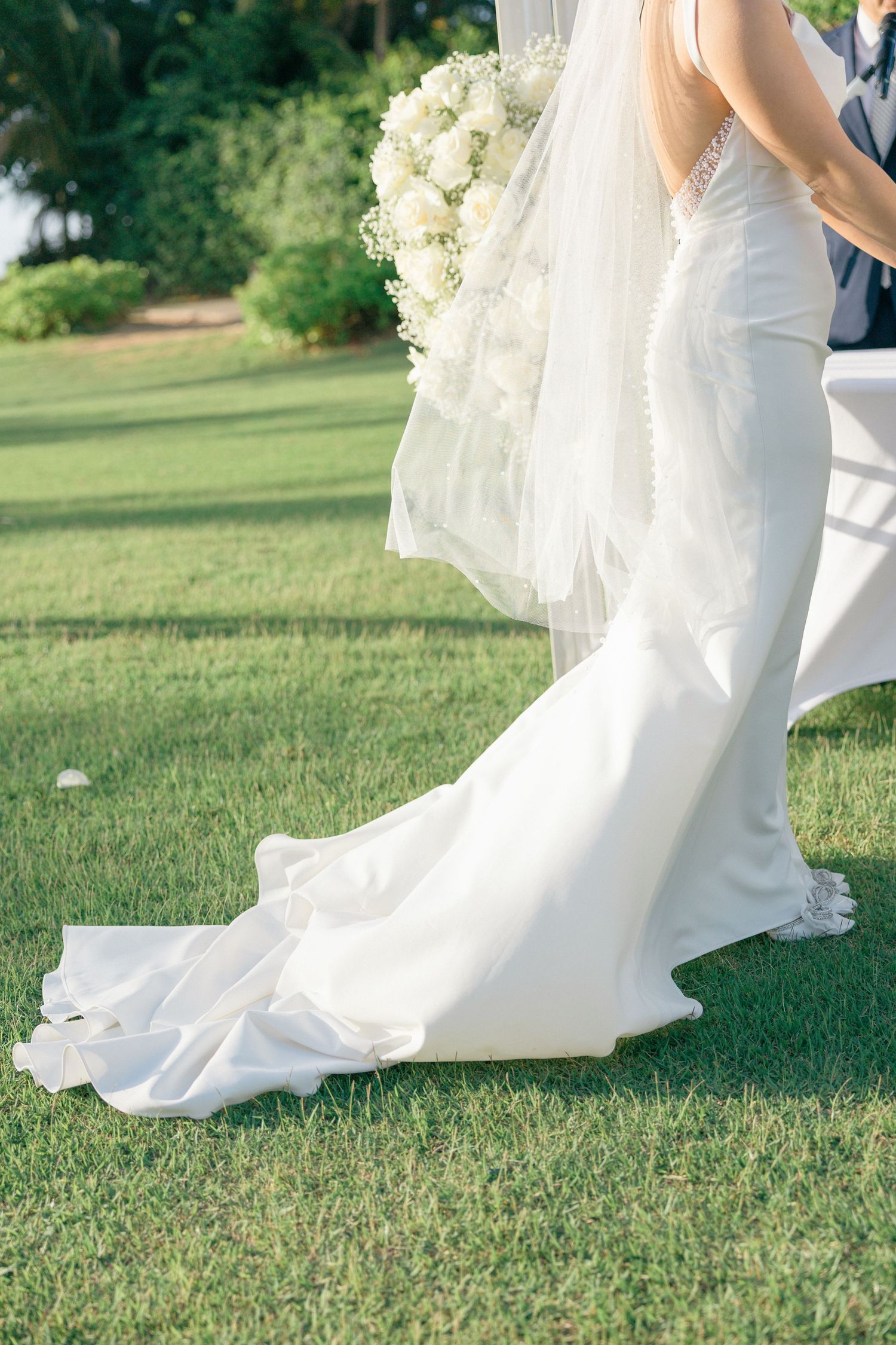 White wedding dress train flowing across green grass during an outdoor ceremony.
