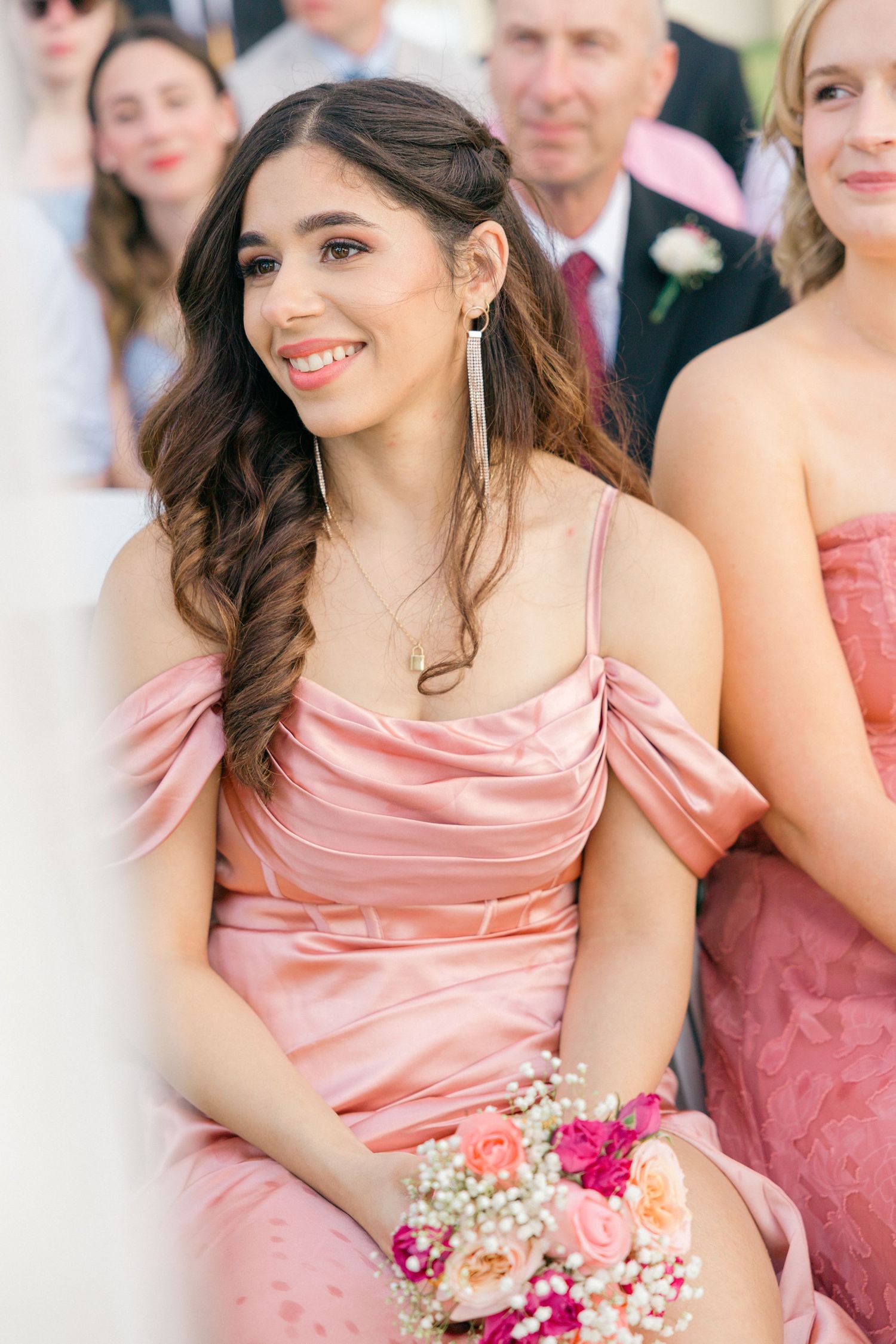 Bridesmaids in coral satin dresses holding pink and white bouquets at outdoor ceremony.