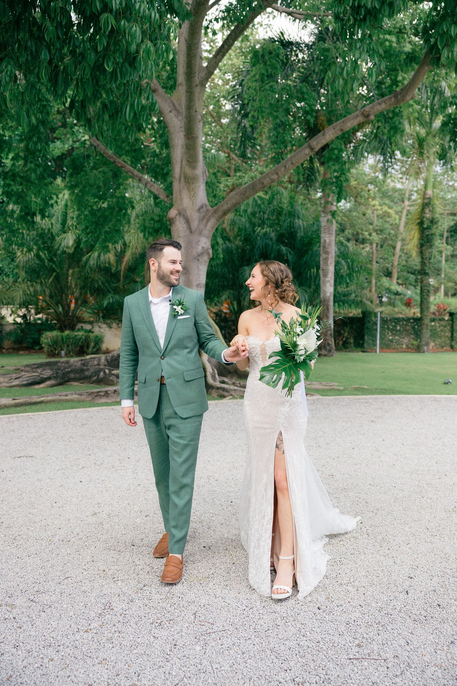 Couple in sage green and white attire walking and laughing together on a gravel path surrounded by trees.