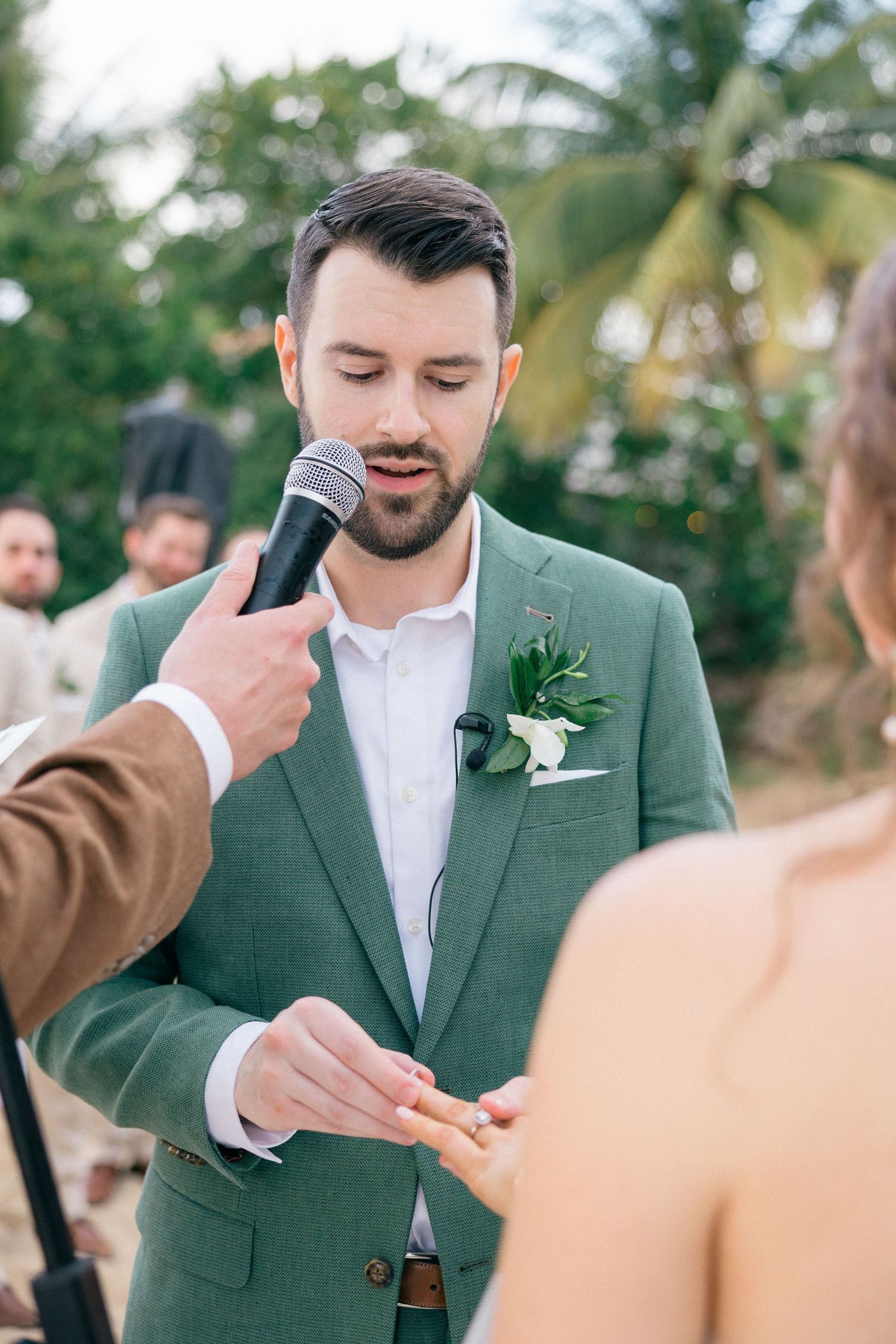 Groom in sage green suit recites vows while holding microphone during beach ceremony.