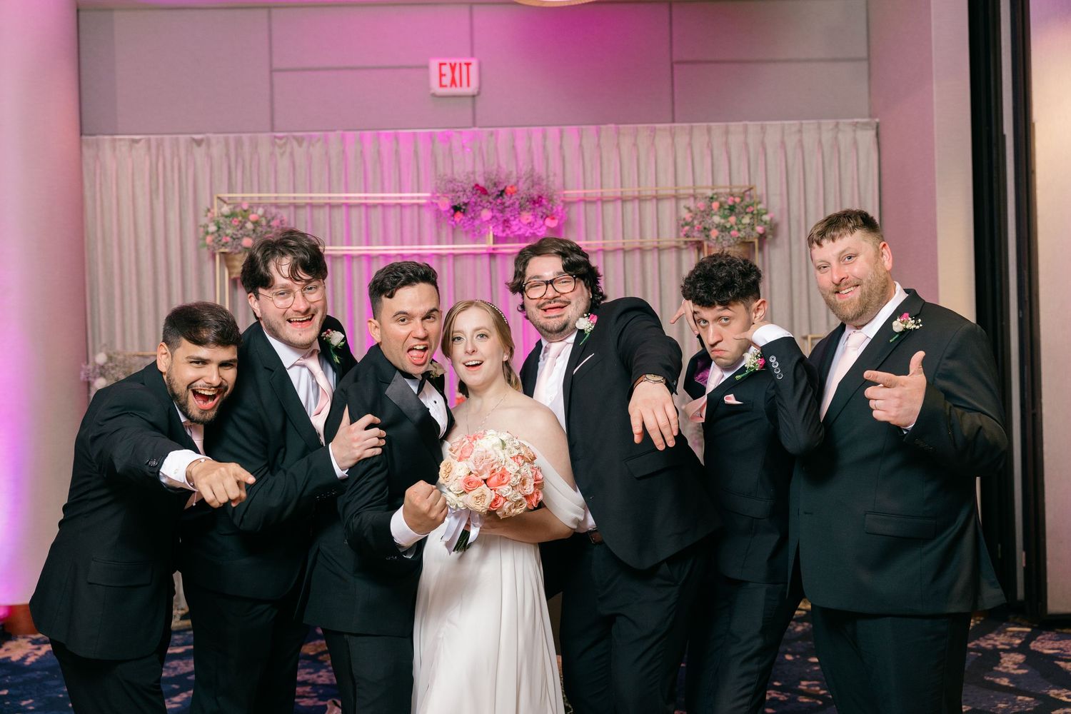 Wedding party in black suits poses playfully with the bride in front of a pink-lit floral backdrop.